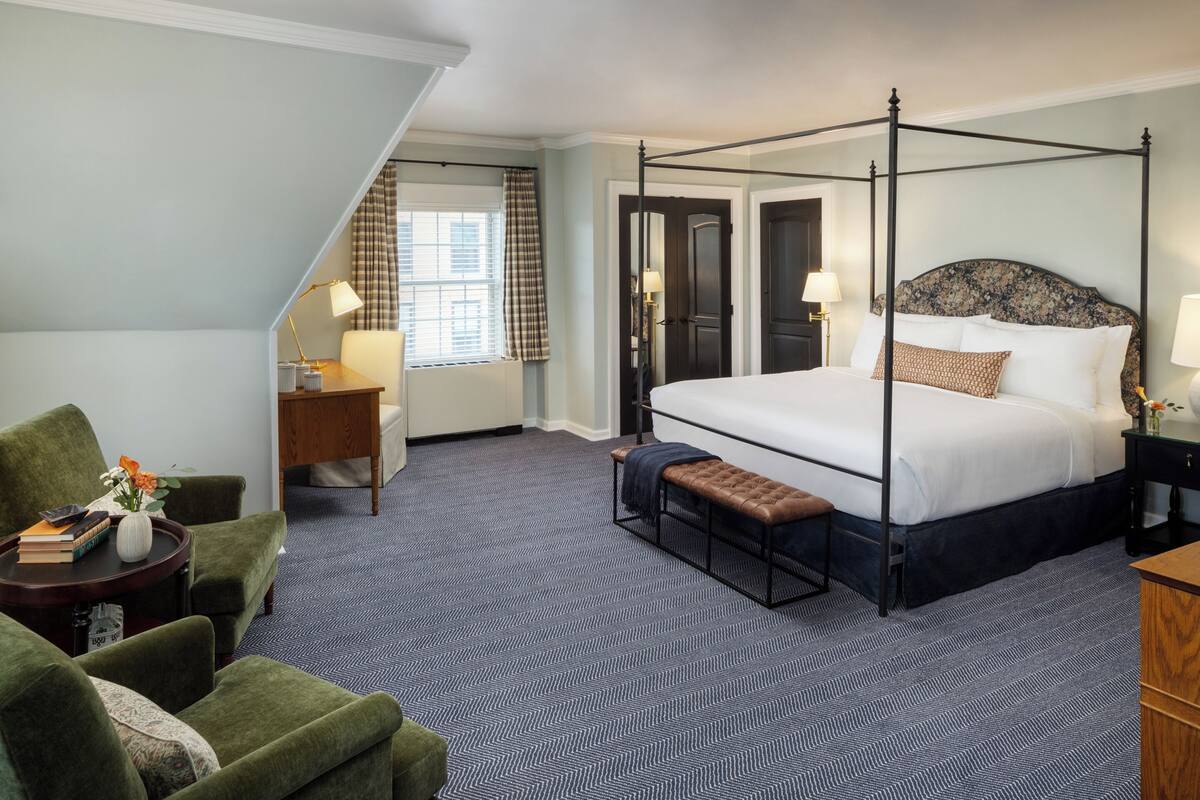 A hotel room with a four-poster bed, a green armchair, a wooden side table, and a bench.