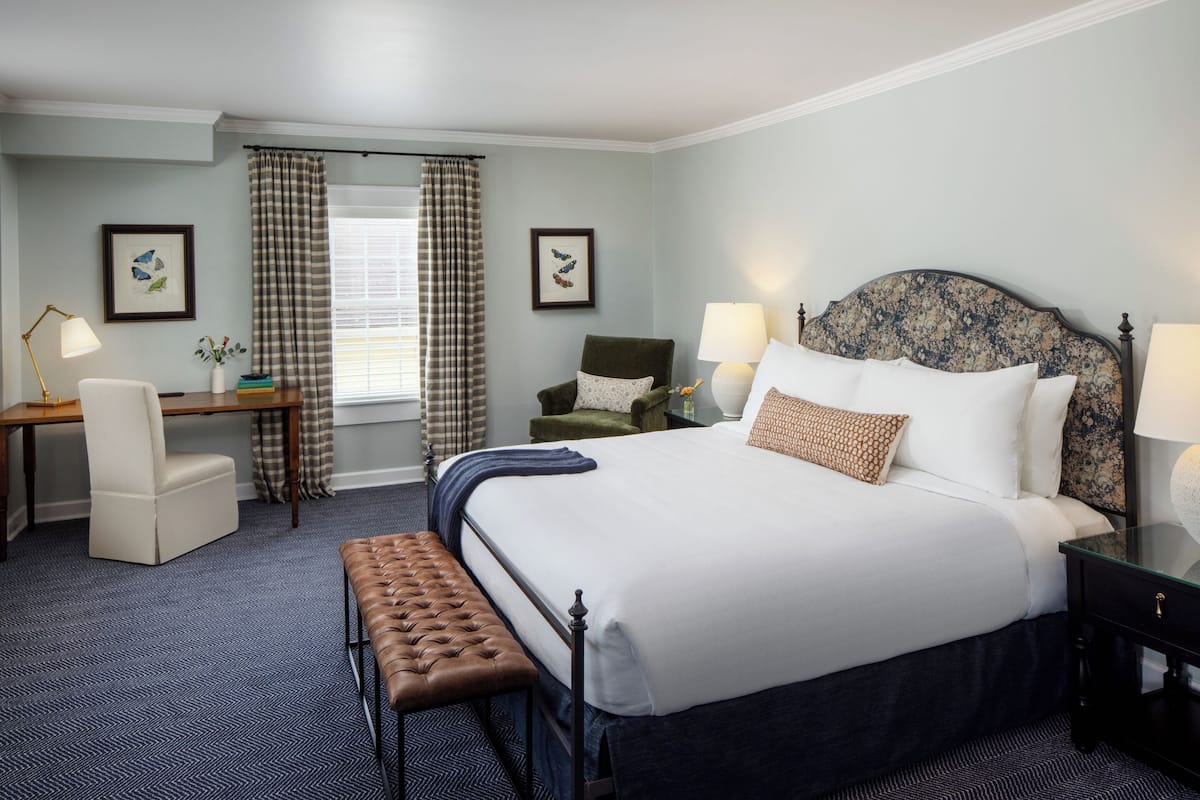 A hotel room with a large bed, a desk, a chair, a bench, and a window with curtains.