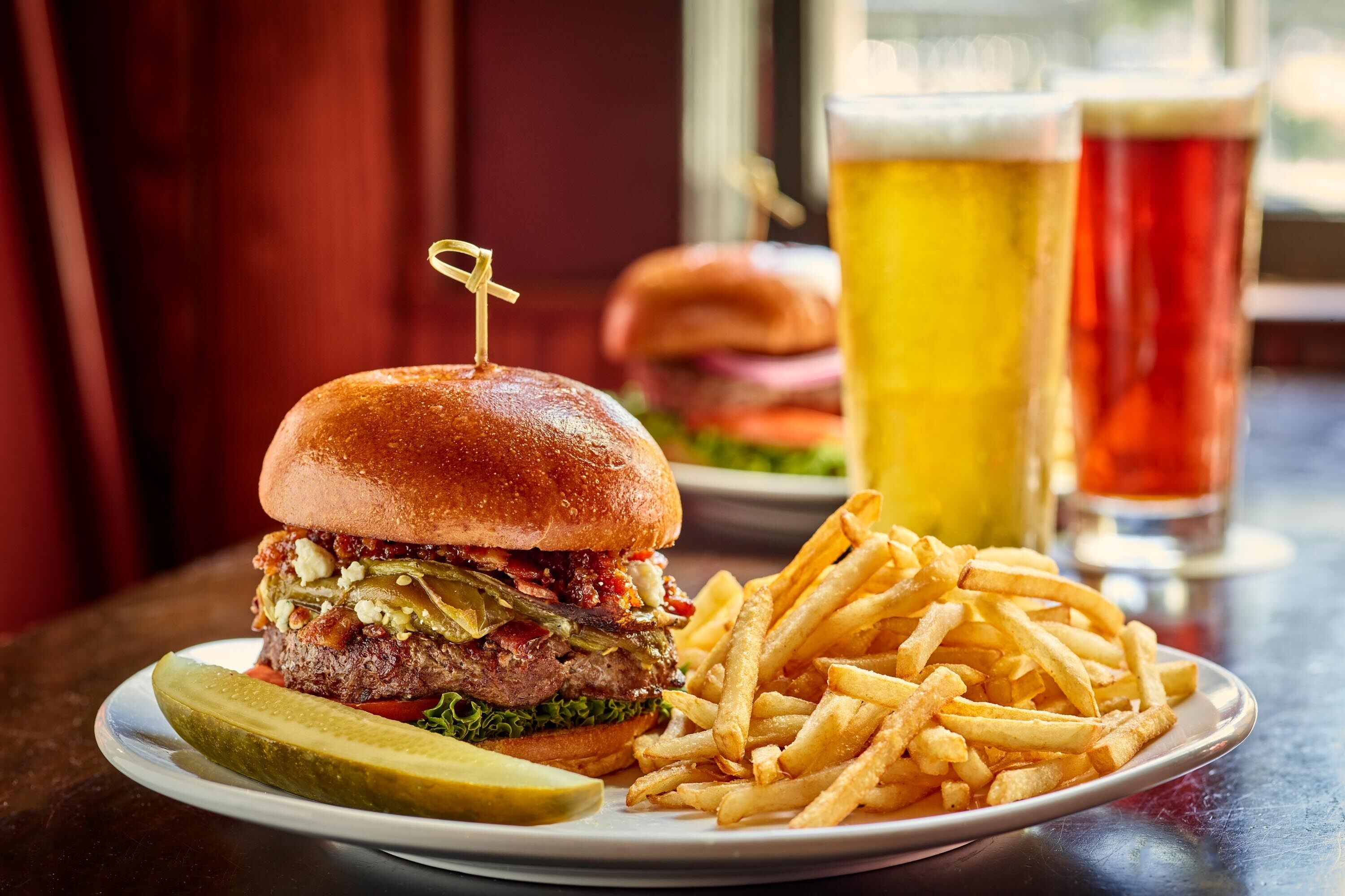 A burger with fries and a pickle on a plate, with a glass of beer in the background.