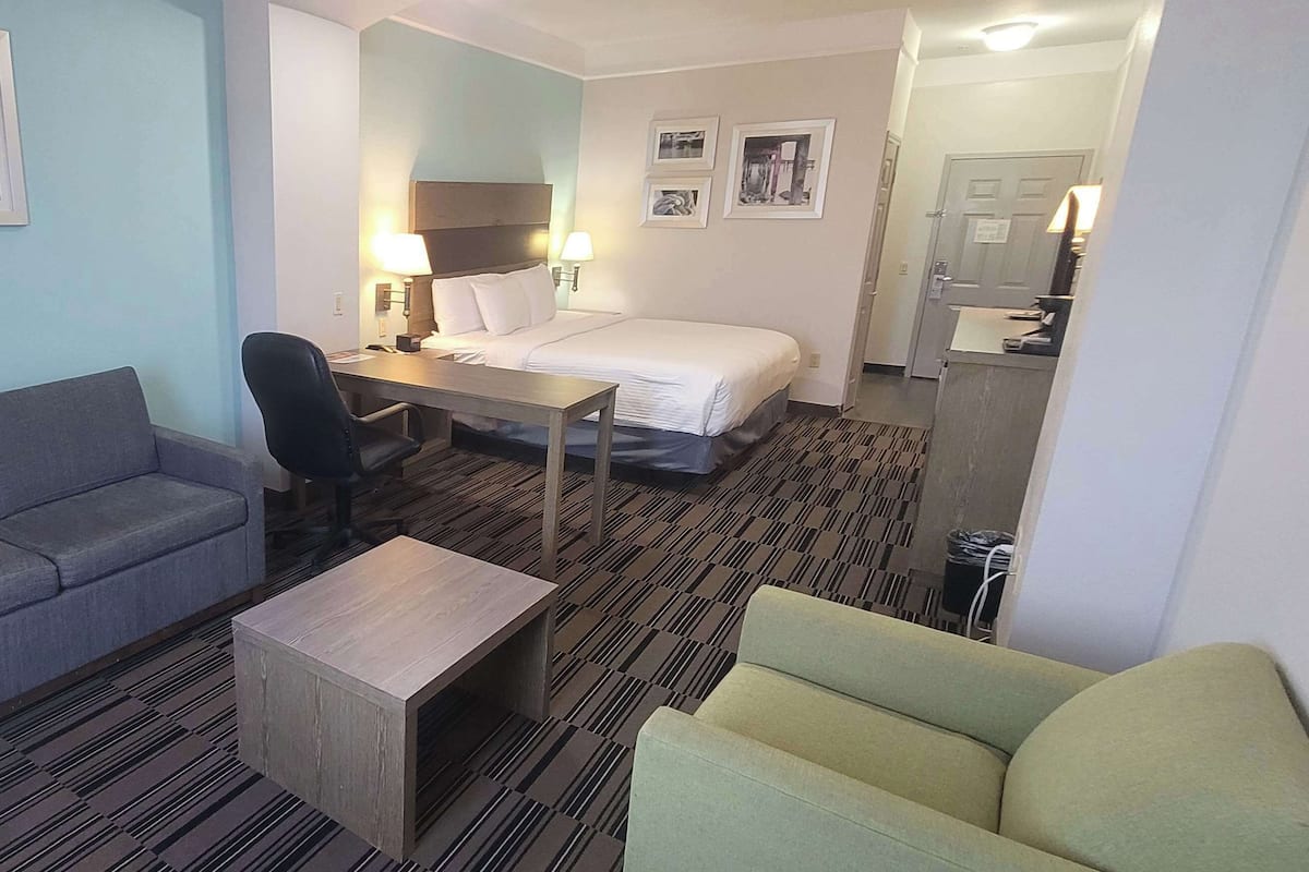 A hotel room with a gray sofa, a small wooden table, a green armchair, a bed with white linens, a desk with a chair, and a mirror above the bed.