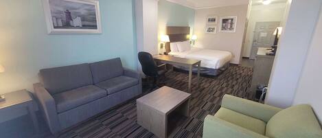 Habitación de hotel con sofá gris, una mesita de madera, un sillón verde, una cama con sábanas blancas, un escritorio con una silla y un espejo sobre la cama.