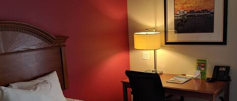 A hotel room with a bed, a desk, a chair, a lamp, and a framed picture on the wall.