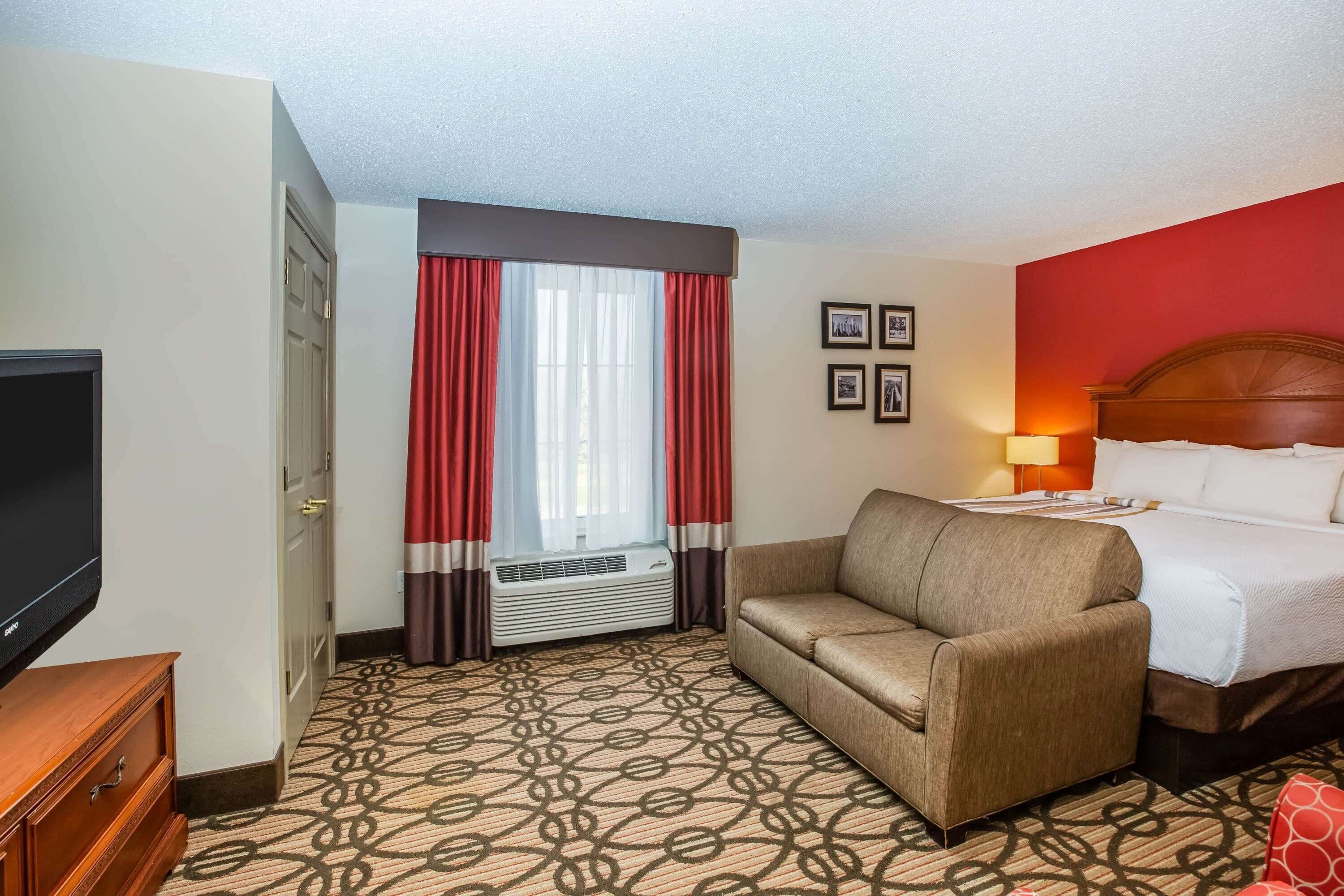 Habitación de hotel con cama, sofá, televisor, ventana con cortinas y alfombra estampada.