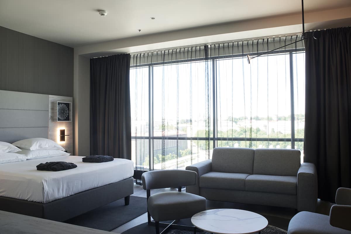 Een moderne hotelkamer met een groot bed, een bank, een tafeltje en uitzicht op de stad door grote ramen.