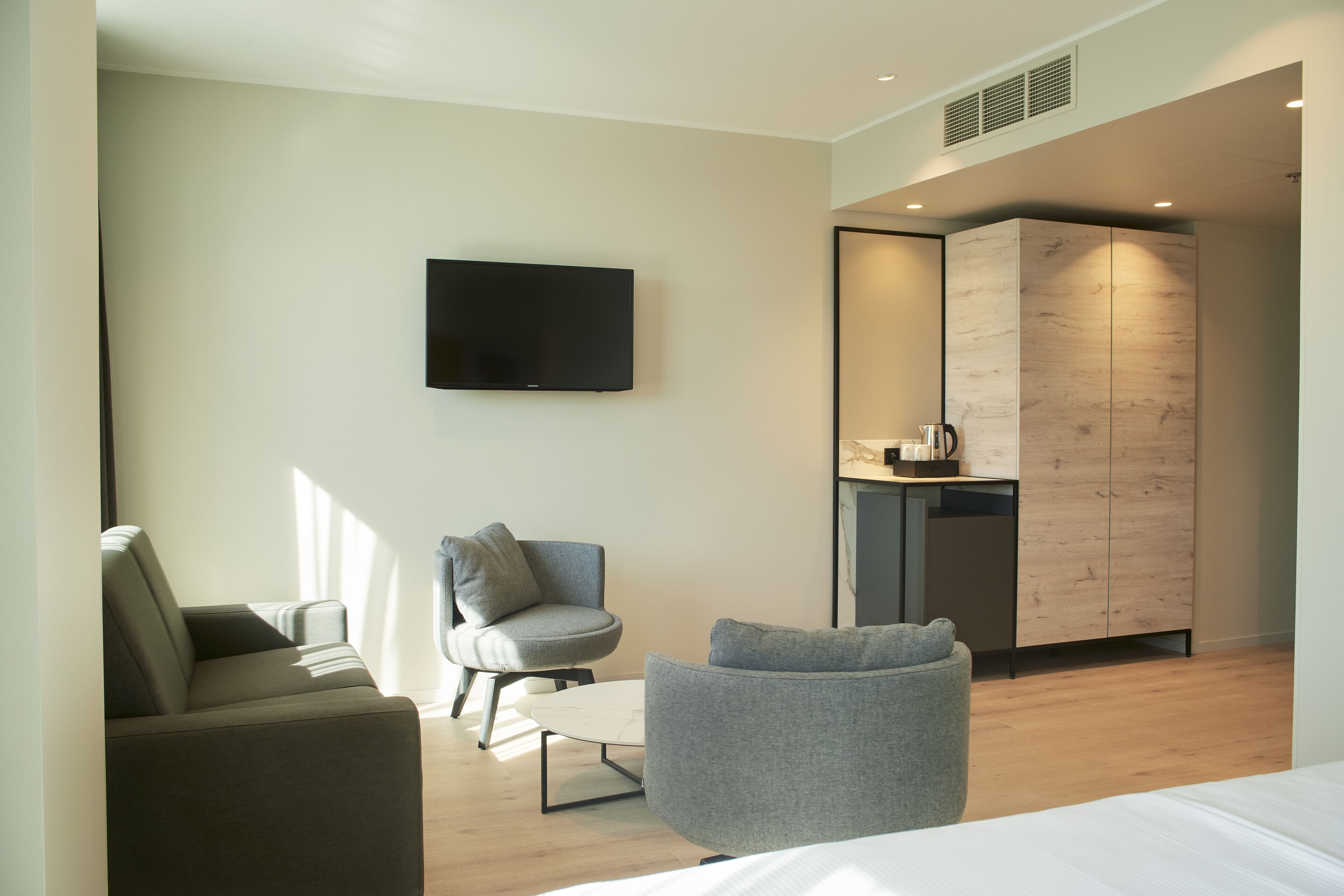 Een moderne hotelkamer met een flatscreen televisie, een grijze bank, een klein rond tafeltje en een houten garderobe.