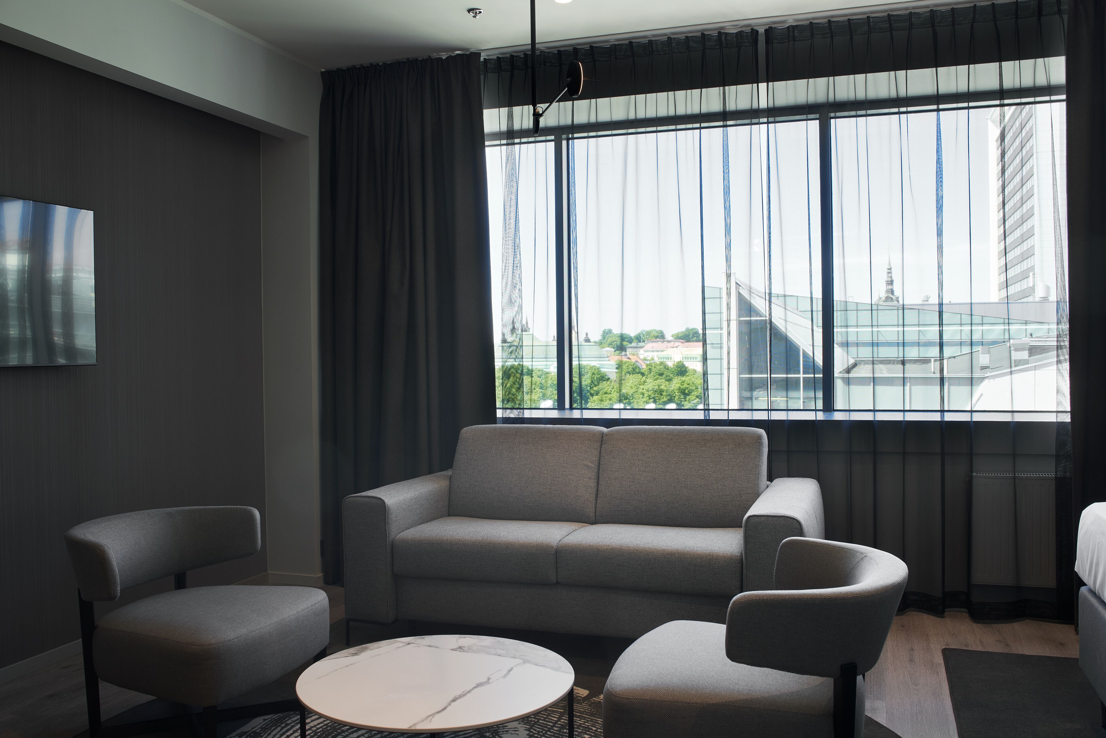 A modern hotel room with a grey sofa, two armchairs, a round coffee table, and a large window offering a city view.