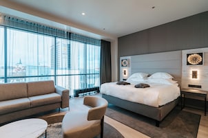 Een moderne hotelkamer met een groot bed, een bank, een stoel en een uitzicht op de stad door grote ramen.