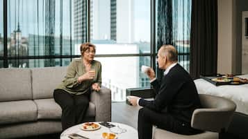 Una habitación de hotel moderna con un ventanal grande, un sofá, una pequeña mesa redonda con alimentos para el desayuno y una cama con colcha blanca.
