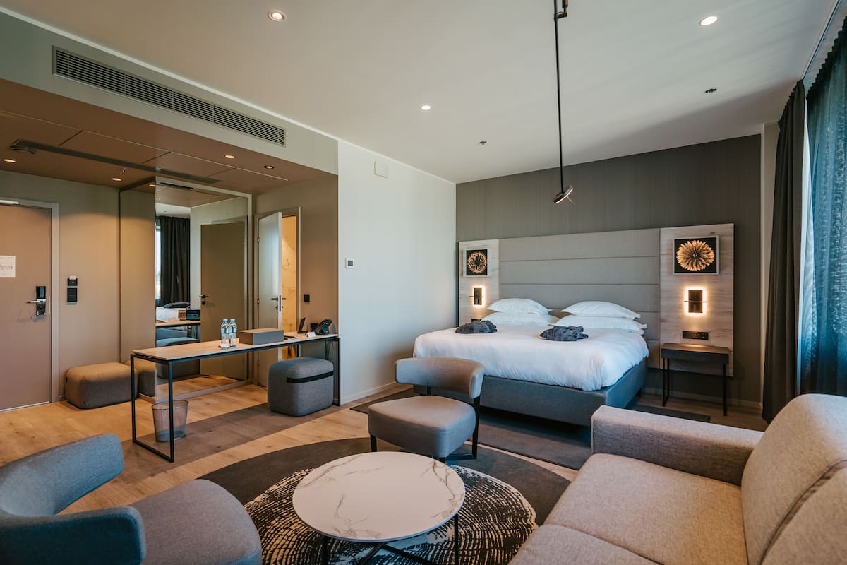Een moderne hotelkamer met een groot bed, een bureau met stoel, een ronde salontafel en een zithoek met banken.