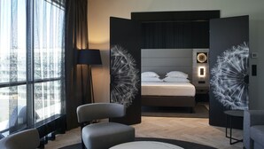 A modern hotel room with a large bed, a seating area with chairs, a round table, and a view of the city through large windows.