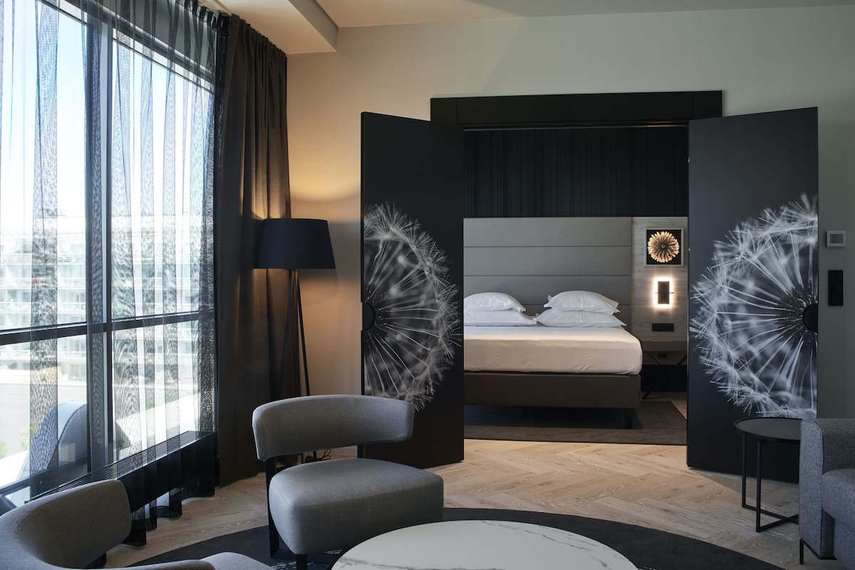Een moderne hotelkamer met een groot bed, een zithoek met stoelen, een ronde tafel en uitzicht op de stad door grote ramen.