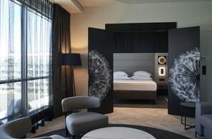 Een moderne hotelkamer met een groot bed, een zithoek met stoelen, een ronde tafel en uitzicht op de stad door grote ramen.