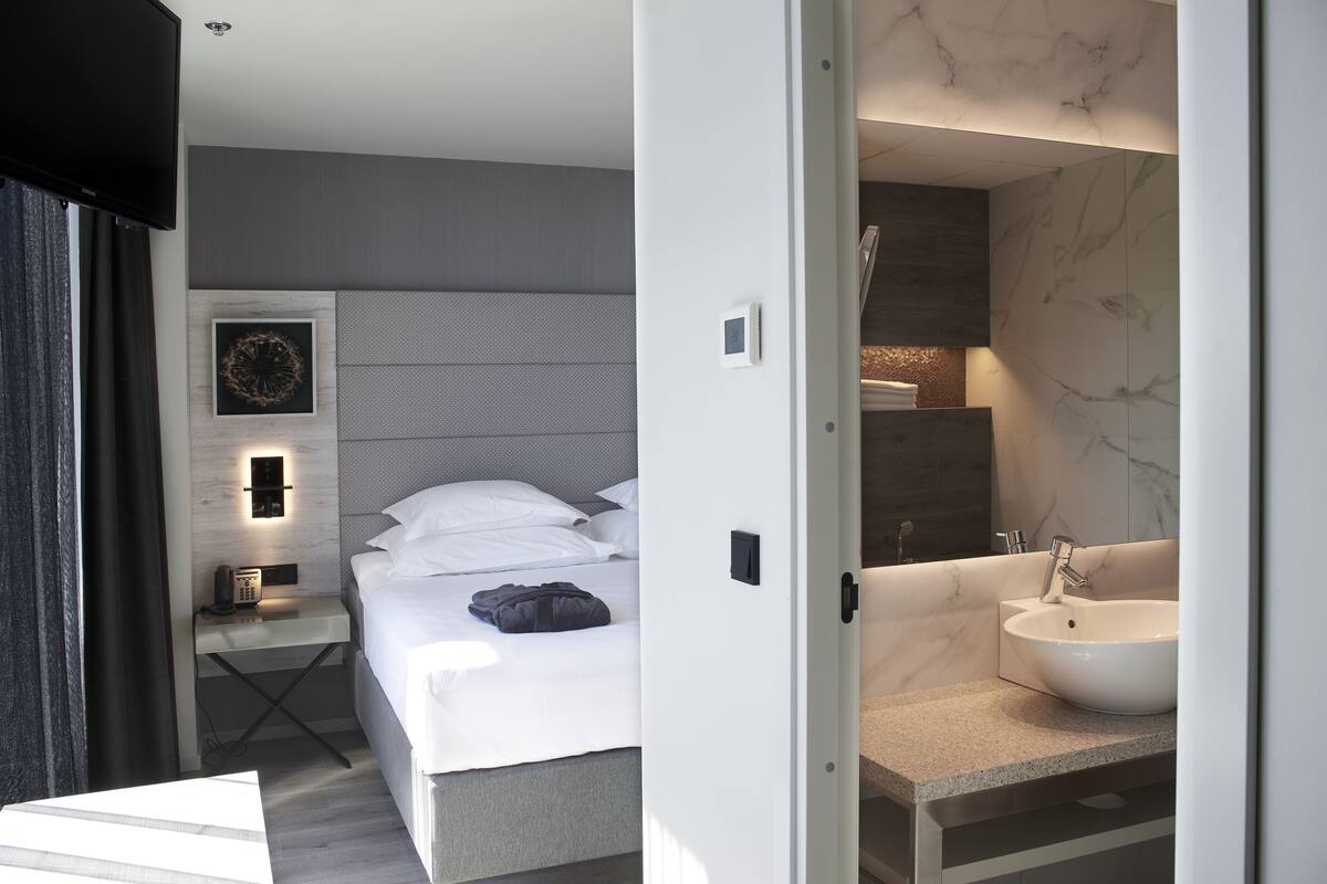 Een moderne hotelkamer met een bed, nachtkastje en een badkamer met wastafel en spiegel.