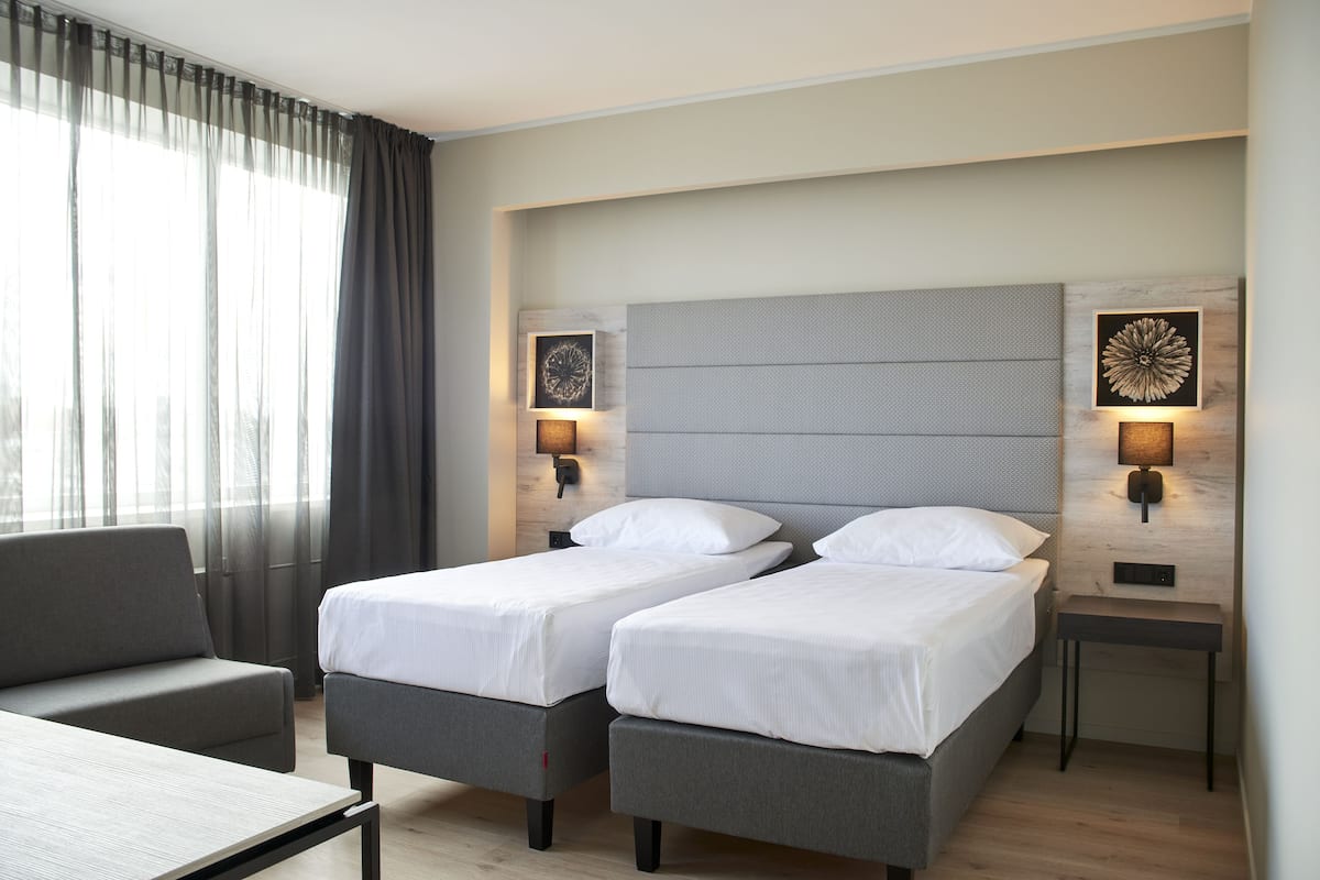 Een hotelkamer met twee eenpersoonsbedden, een slaapbank, een tafel en een groot raam met gordijnen.