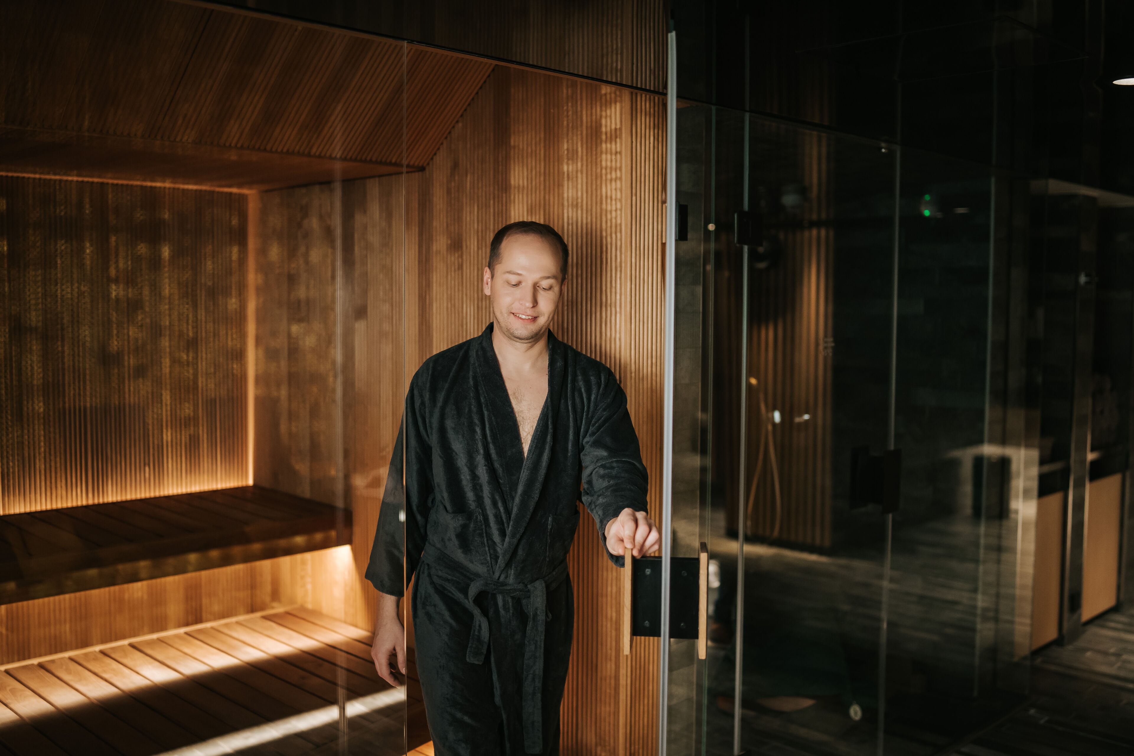A person in a black robe standing in a modern, dimly lit room with wooden paneling and a glass door.