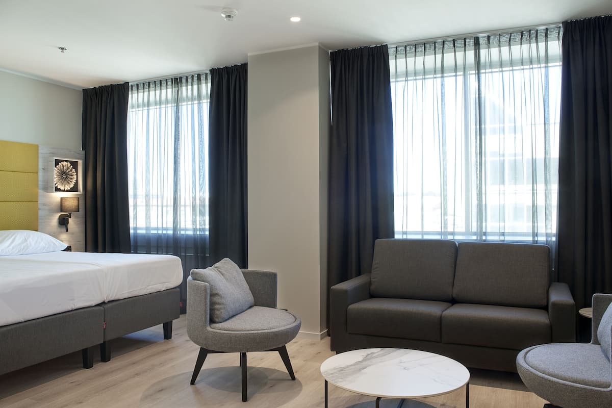 Een hotelkamer met een bed, twee banken, een ronde salontafel en grote ramen met gordijnen.