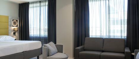 Una habitación de hotel con una cama, dos sofás, una mesa de centro redonda y ventanas grandes con cortinas.