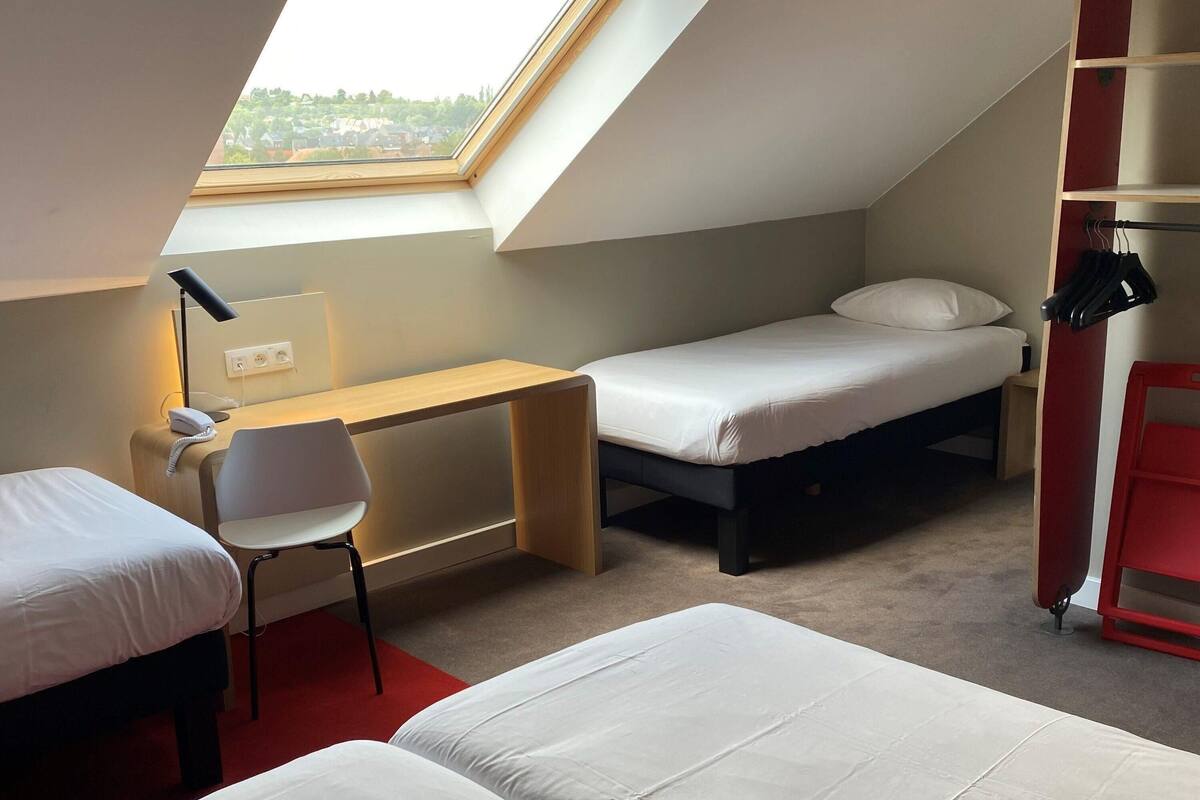 Habitación pequeña con tragaluz, escritorio con silla, dos camas individuales y un mueble de almacenamiento rojo.