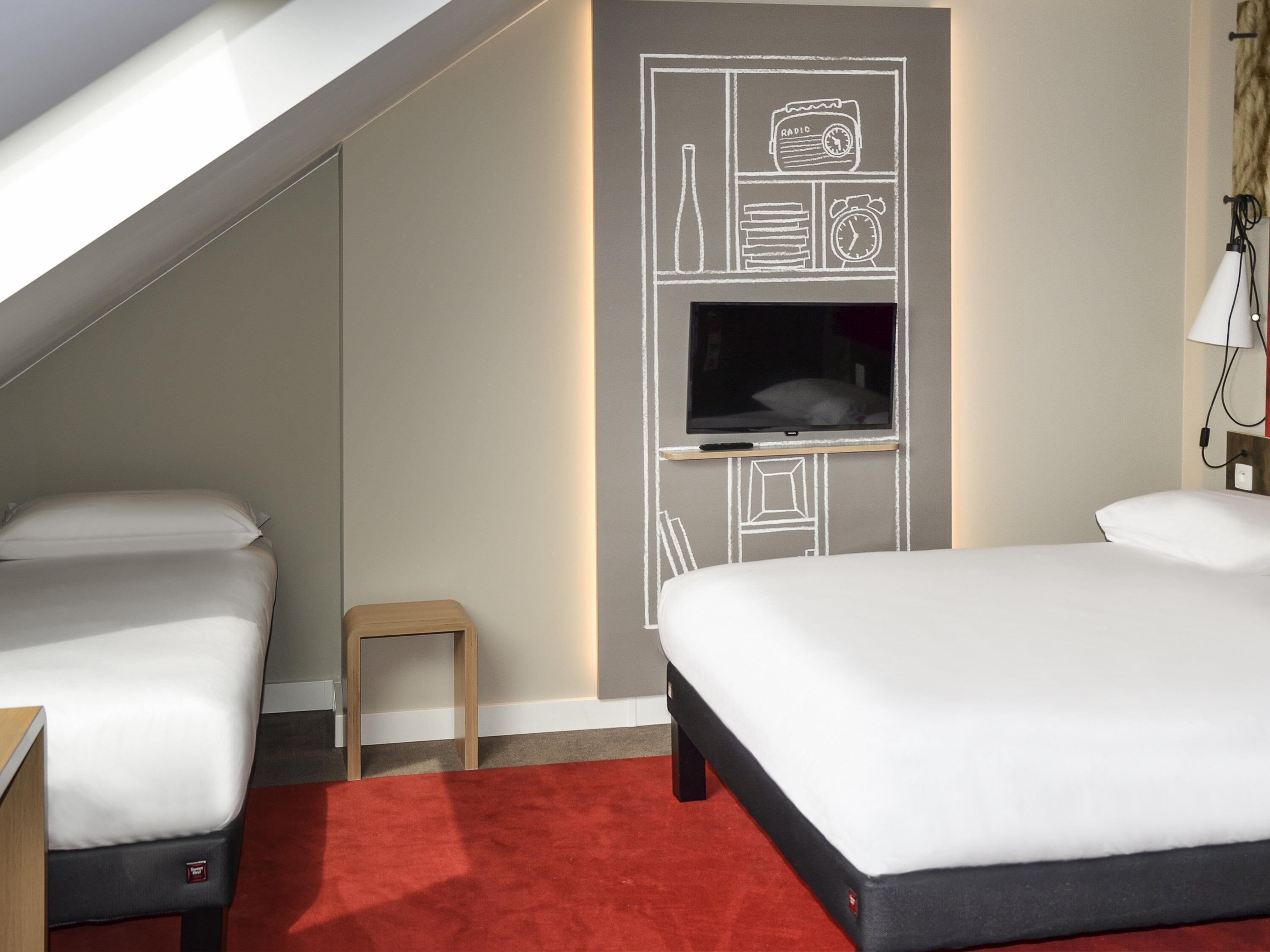 A hotel room with two beds, a TV mounted on the wall, and a small wooden stool.