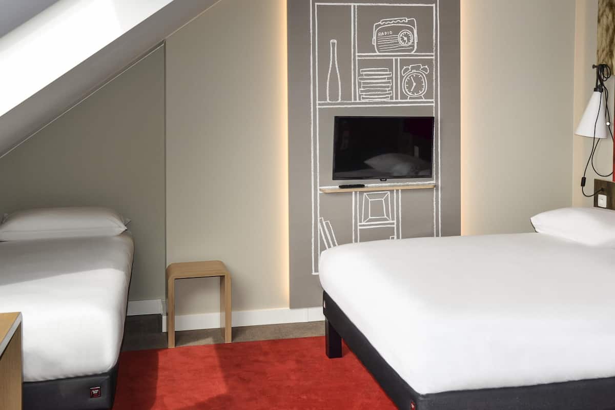 A hotel room with two beds, a TV mounted on the wall, and a small wooden stool.