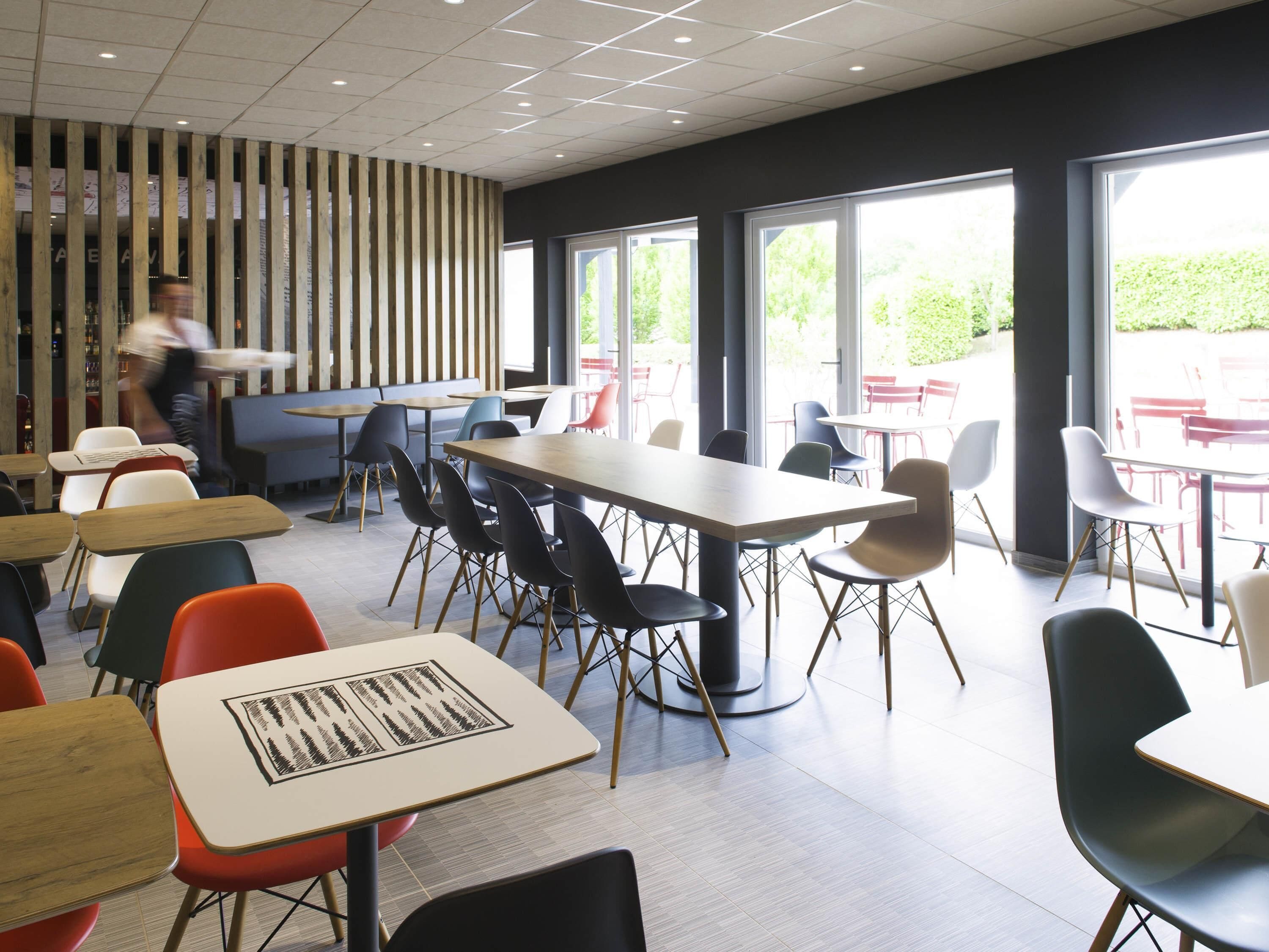 A modern dining area with multiple tables and chairs, large windows, and a wooden slat wall.