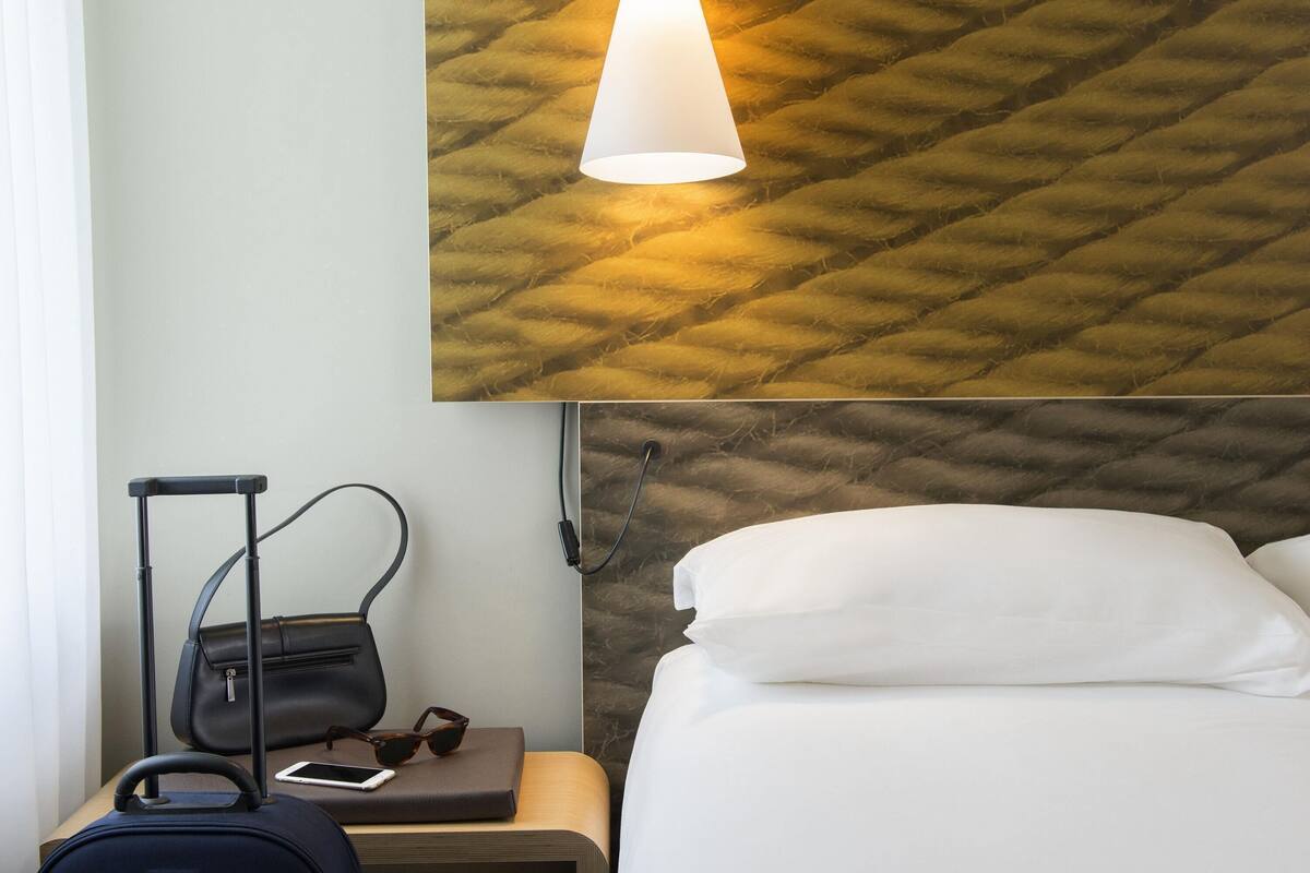 Una habitación de hotel con una cama, una mesita de noche, una maleta, un bolso de mano, un par de gafas de sol y una lámpara de pared.