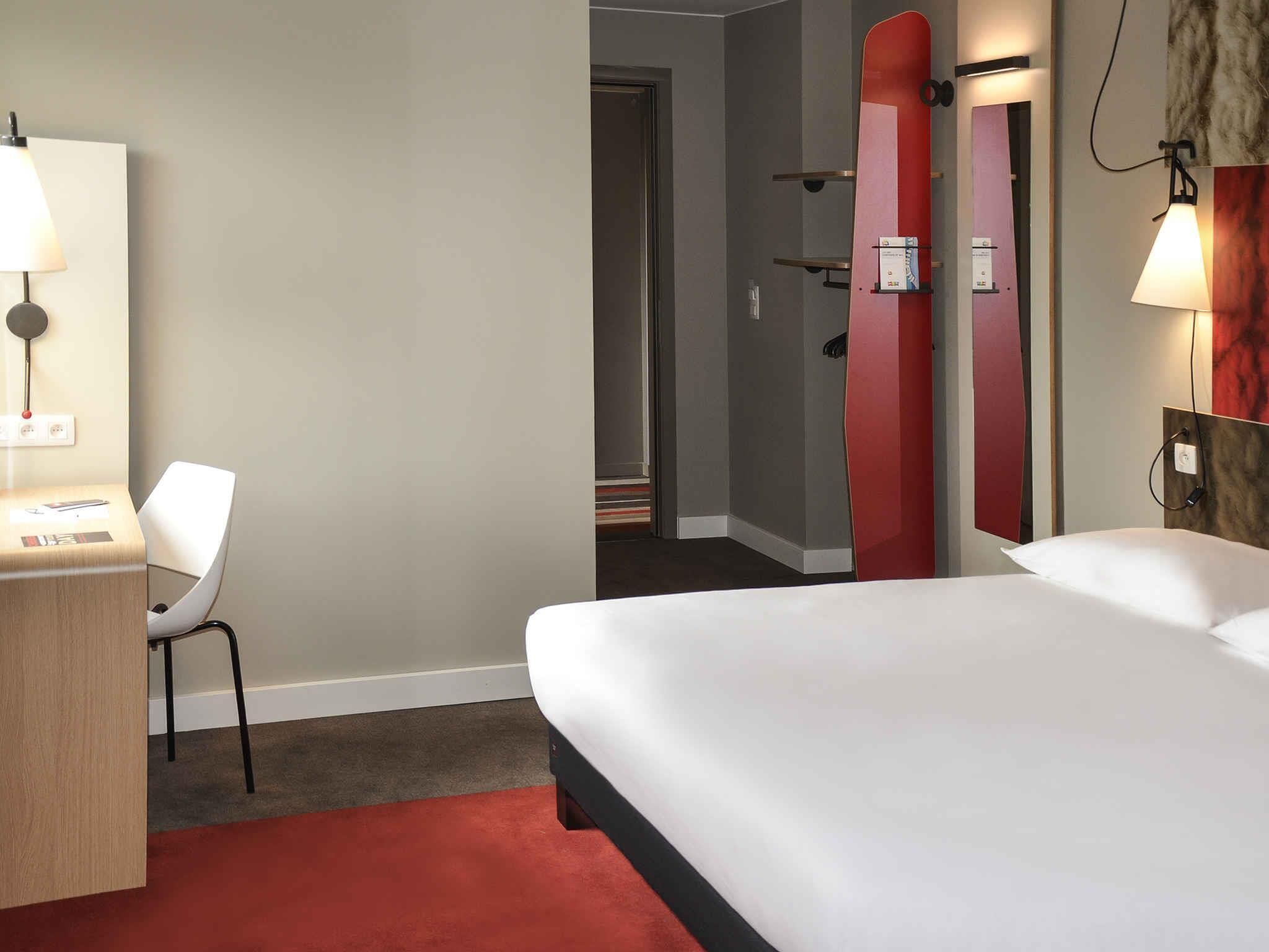 A hotel room with a bed, a desk, a chair, a mirror, and a red carpet.