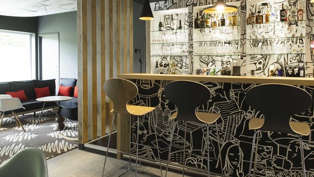 Una zona de bar con mostrador de madera, sillas negras y una pared con botellas y vasos para bebidas.