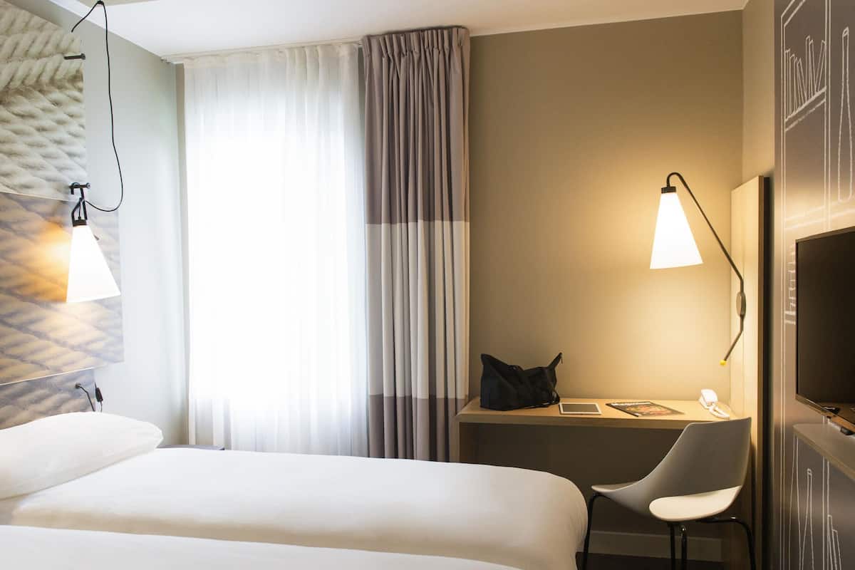 Una habitación de hotel con una cama, un escritorio con silla, un televisor y una lámpara.