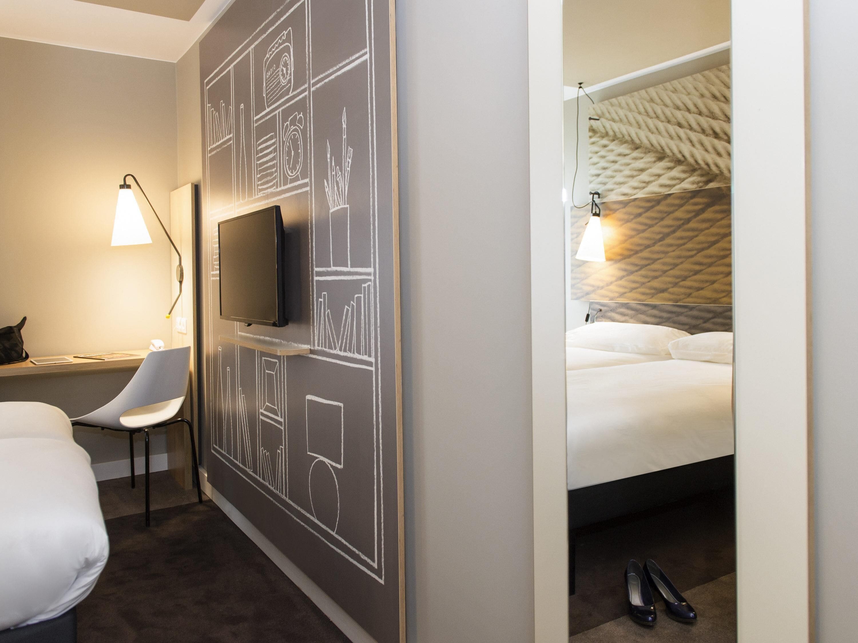 A hotel room with a bed, a desk with a chair, a television, and a wall with chalkboard drawings.