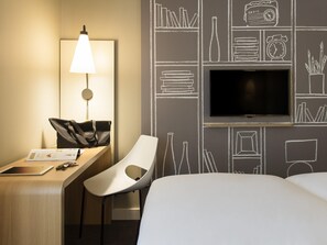 A hotel room with a desk, chair, lamp, TV, and a wall with chalkboard-style drawings.