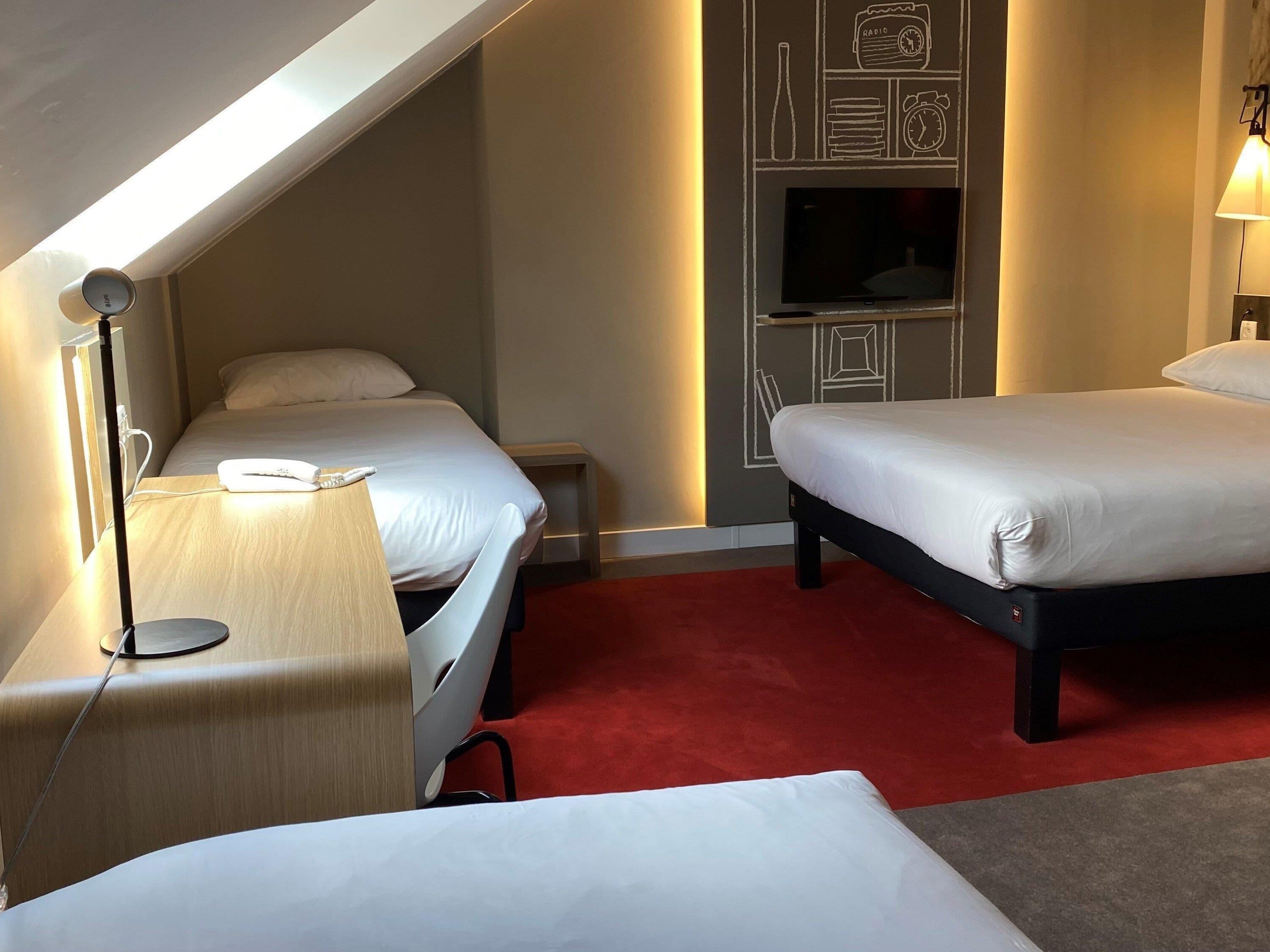 Una habitación de hotel con dos camas, un escritorio con lámpara, un televisor y una alfombra roja.