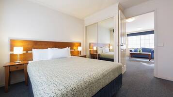 A hotel room with a large bed, bedside tables with lamps, a mirror, and a view of a blue sofa and window in the adjacent room.