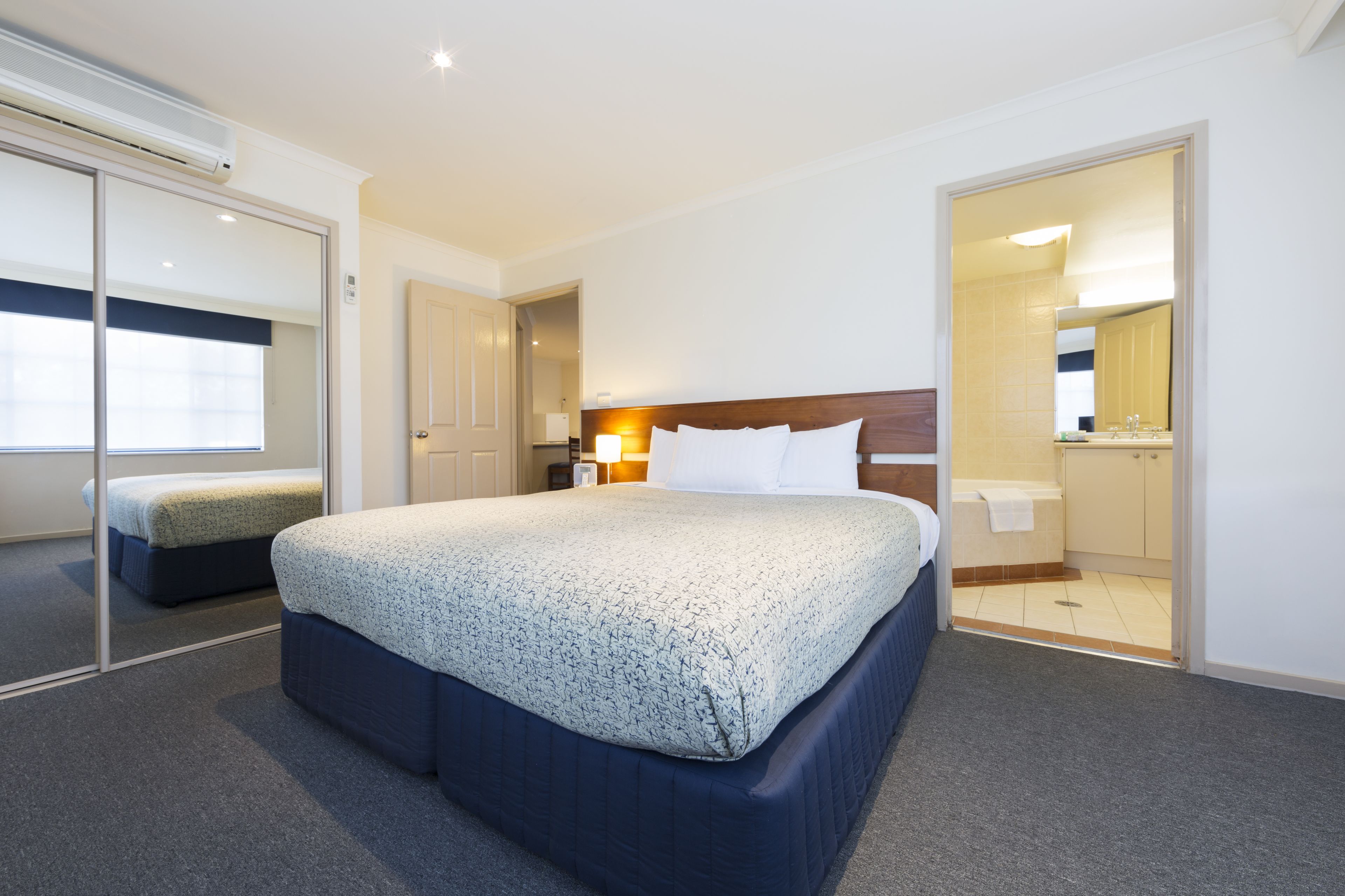 A hotel room with a large bed, a bedside table with a lamp, a bathroom visible through an open door, and a sliding glass door leading to a balcony.