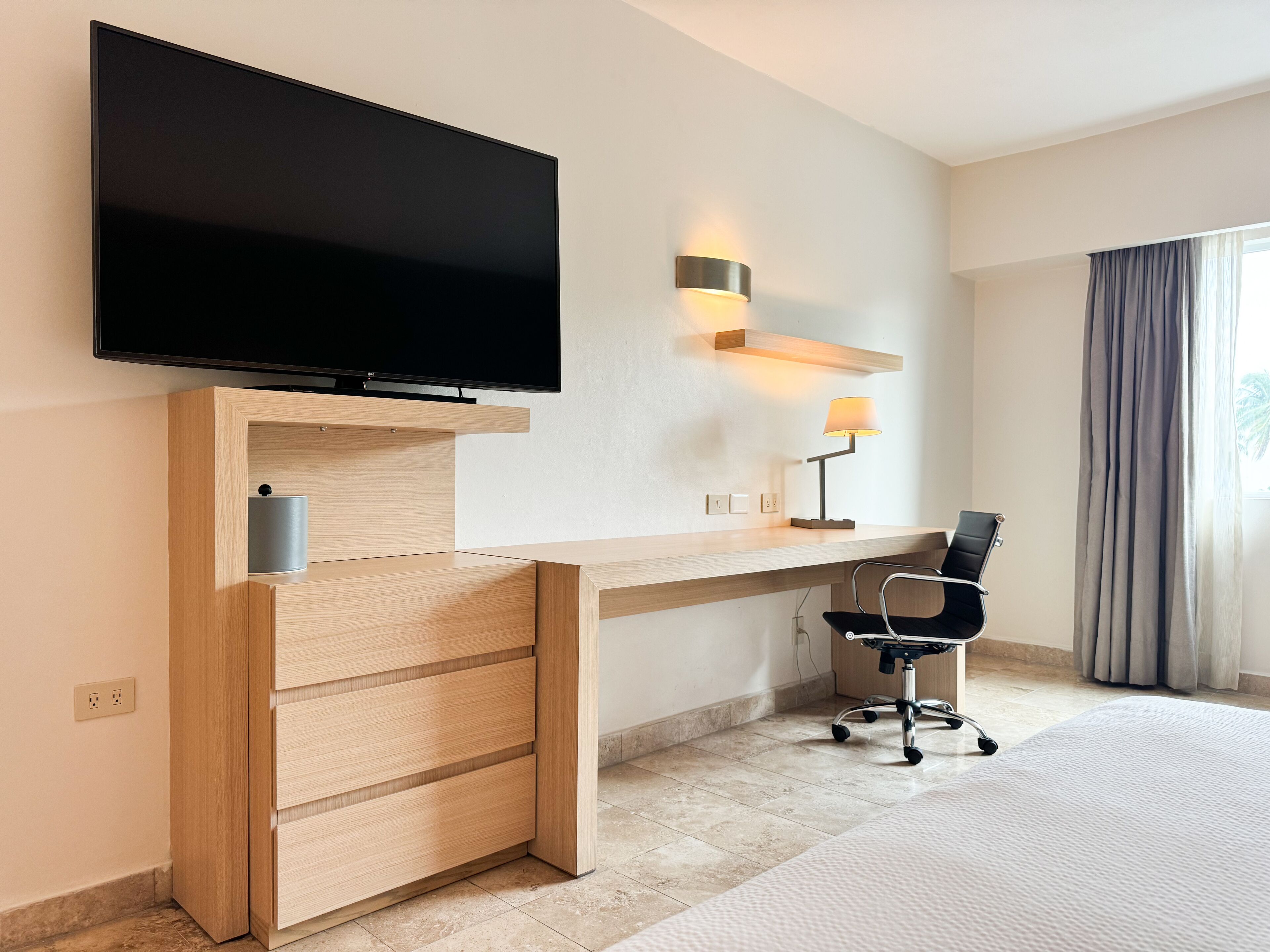 Habitación de hotel moderna con televisión de pantalla plana montada sobre un escritorio de madera, una lámpara de escritorio, una silla y un ventanal con cortinas.