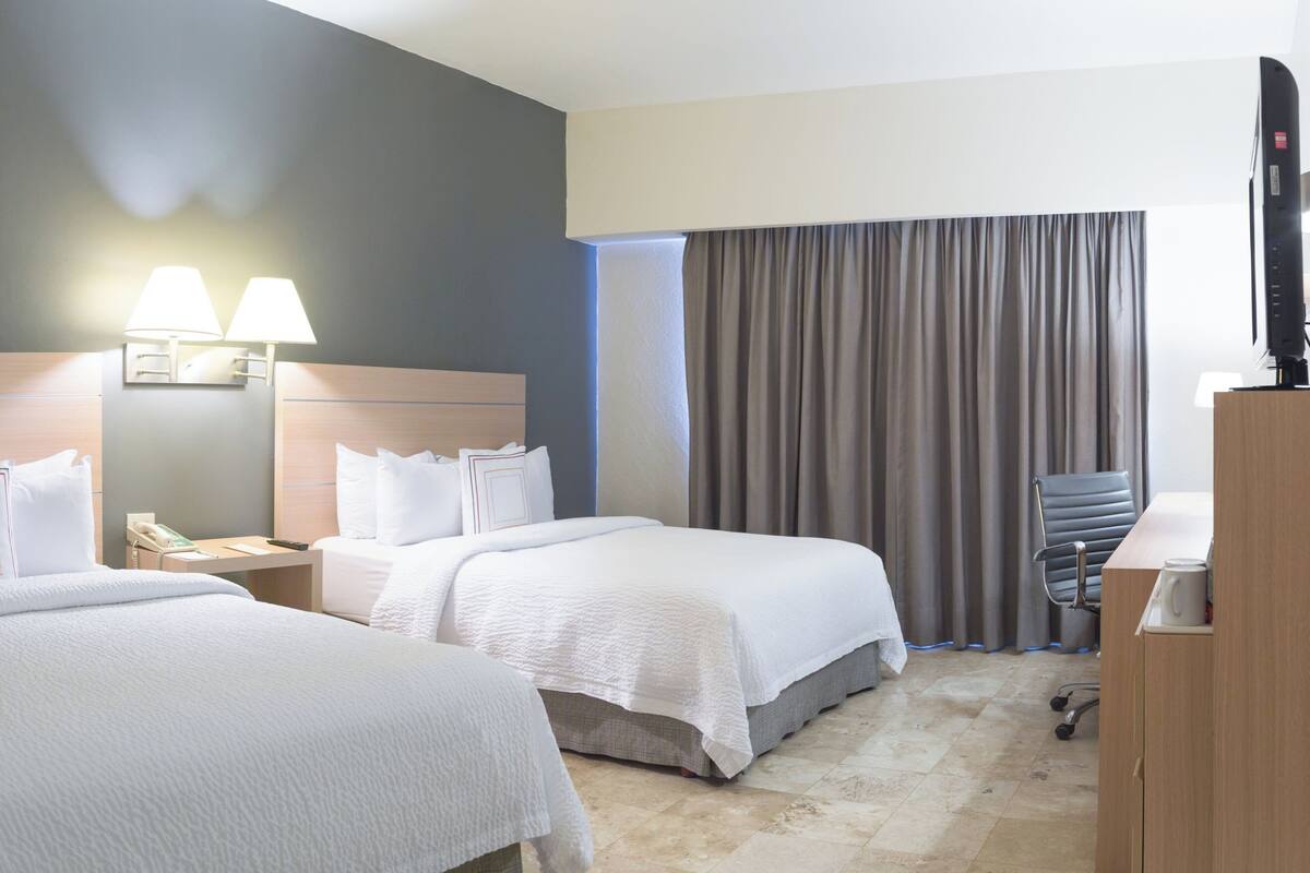A hotel room with two beds, a desk, a chair, a television, and a window with curtains.