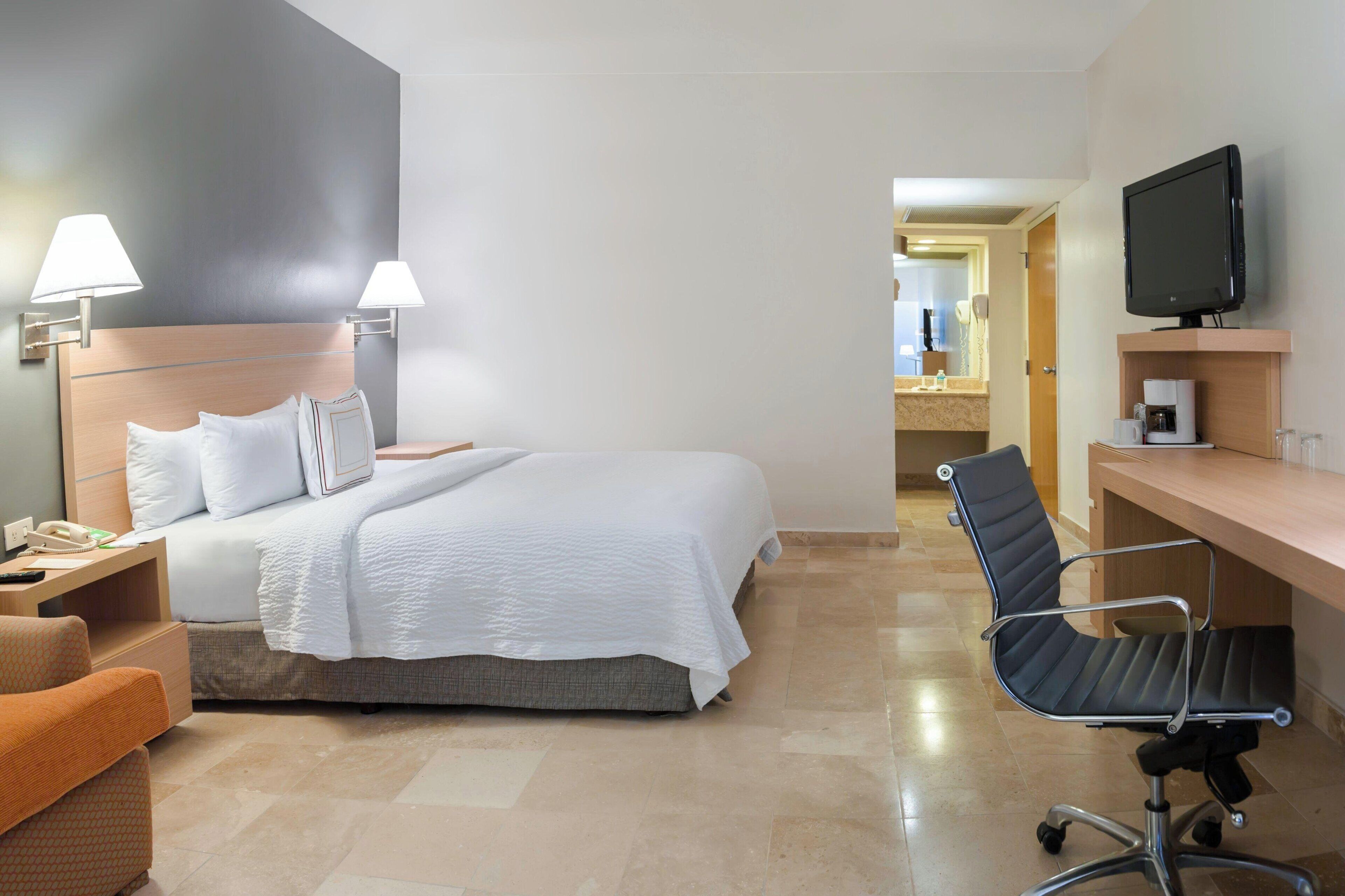A hotel room with a bed, bedside lamps, a desk with a chair, a television, and a bathroom visible through an open door.