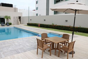 Una piscina con suelo de baldosas, una mesa con sillas y un paraguas grande.