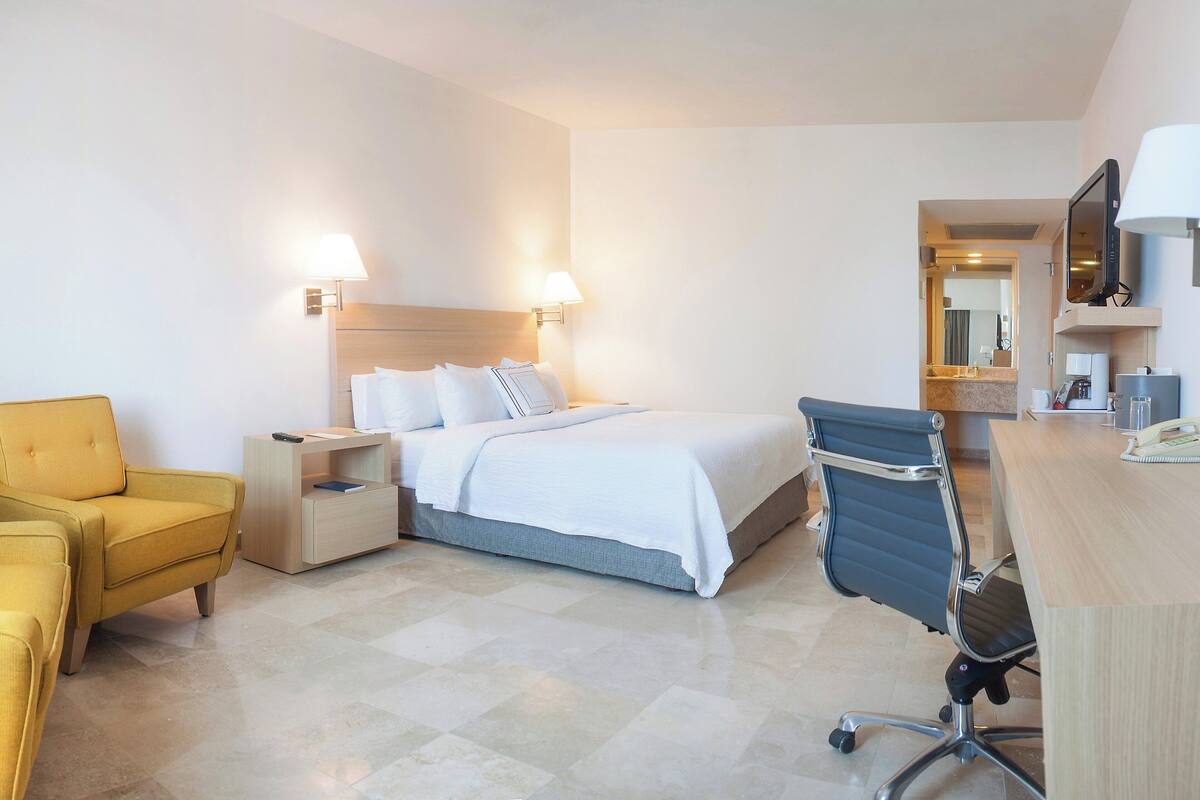 A hotel room with a bed, a desk, a chair, a nightstand, and a lamp.
