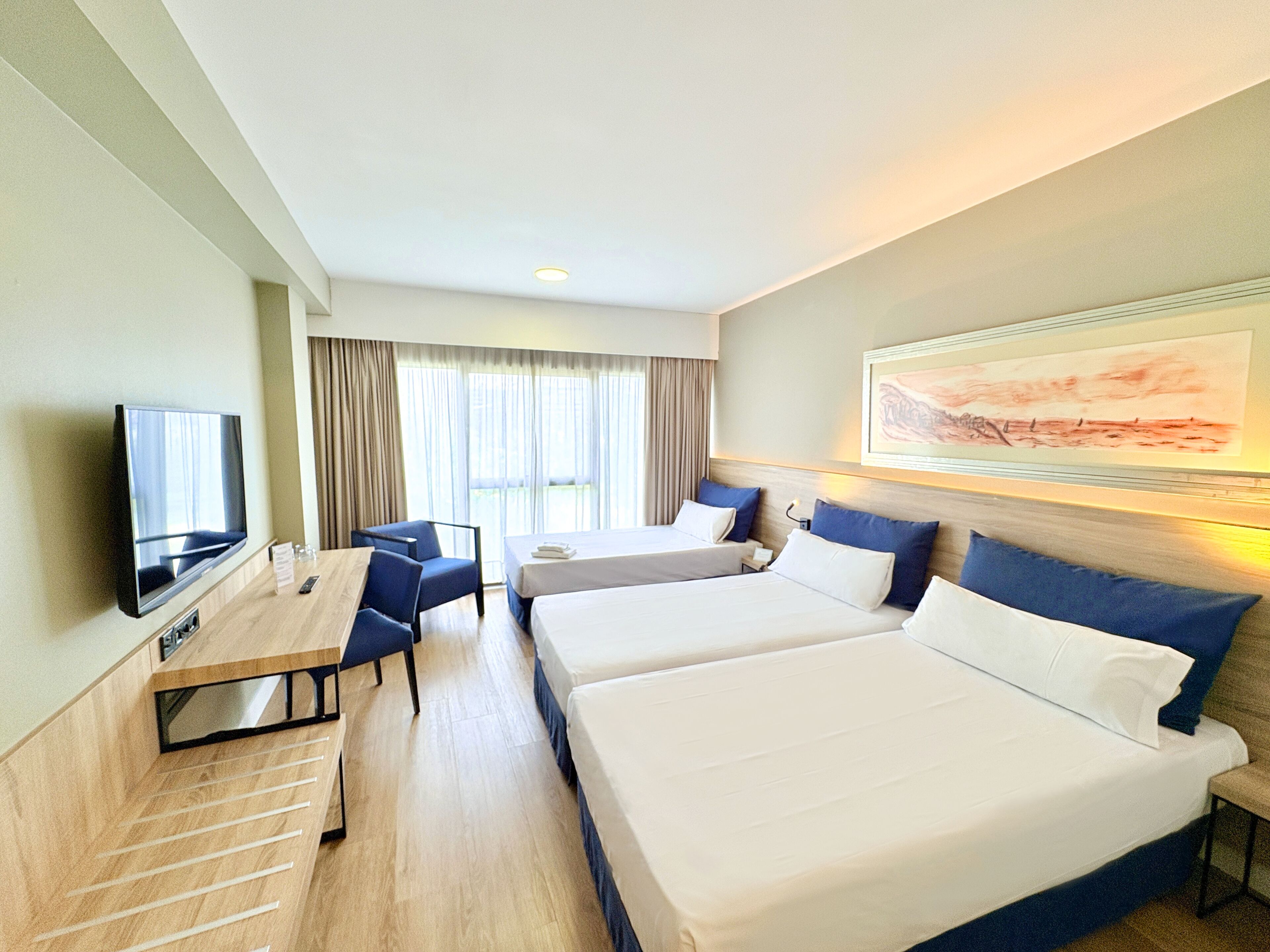 A hotel room with two beds, a desk with a chair, a TV, and a large window with curtains.