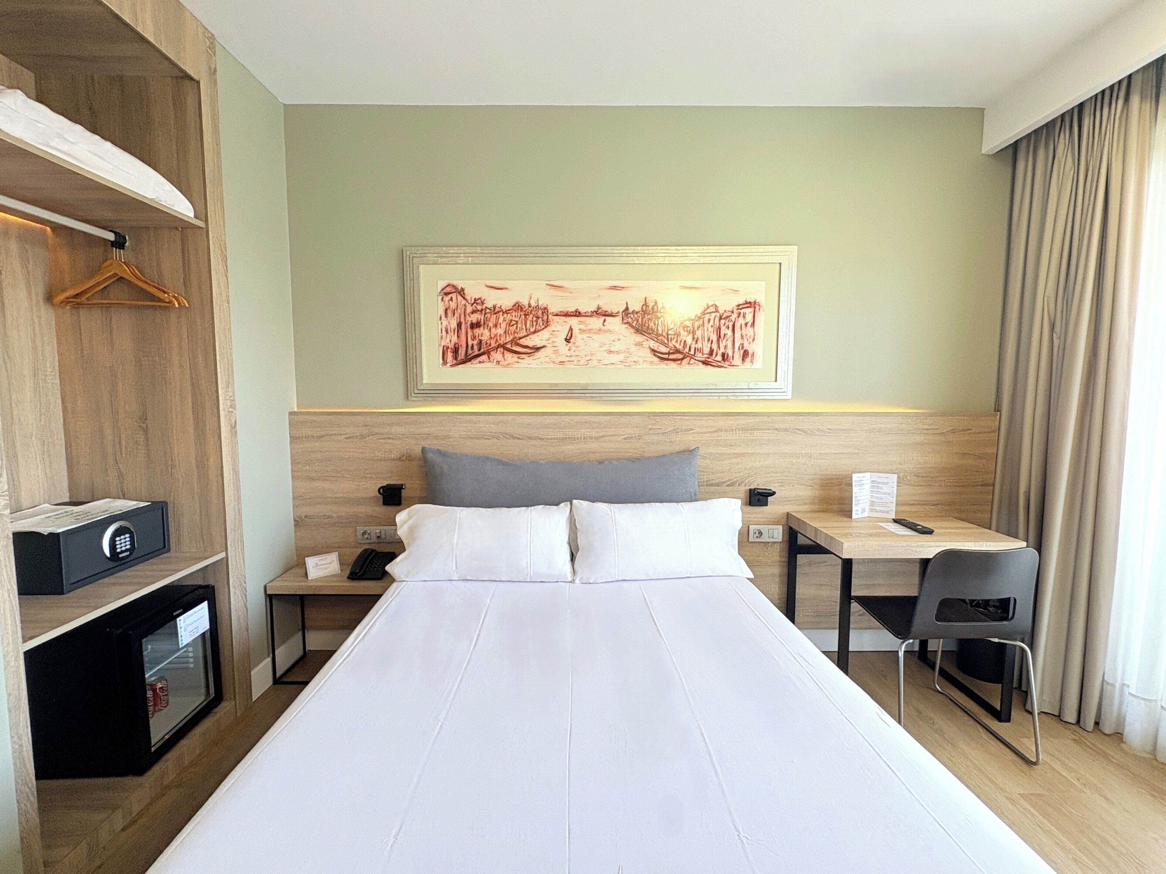 Ein Hotelzimmer mit einem großen Bett, einem Schreibtisch mit Stuhl, einem kleinen Safe, einem Telefon und einem gerahmten Bild an der Wand.