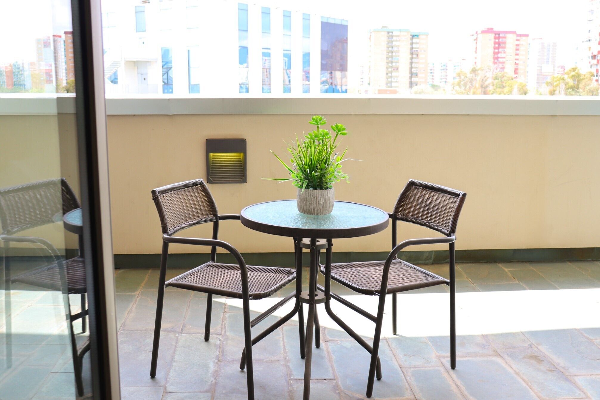 A balcony with a round table and four wicker chairs, a potted plant, and a wall-mounted heater.