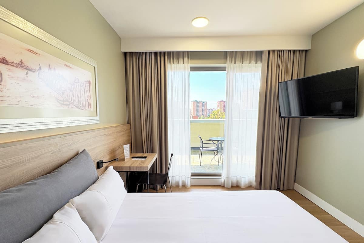 Een hotelkamer met een groot bed, een bureau met stoel, een aan de muur bevestigde televisie en uitzicht op de stad door het raam.