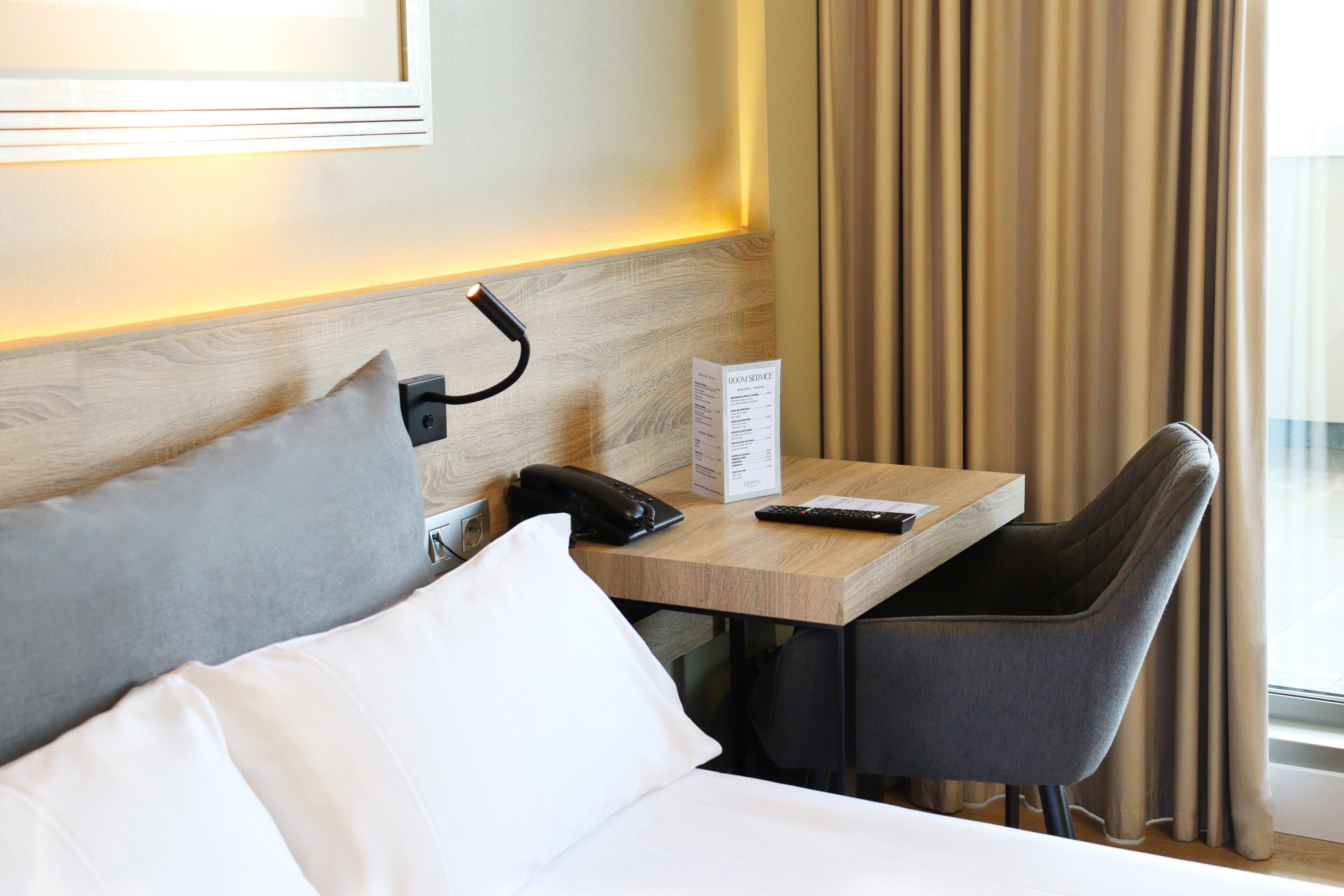 Ein Hotelzimmer mit Bett, Schreibtisch, Telefon, Fernbedienung, Stuhl und einem Fenster mit Vorhängen.