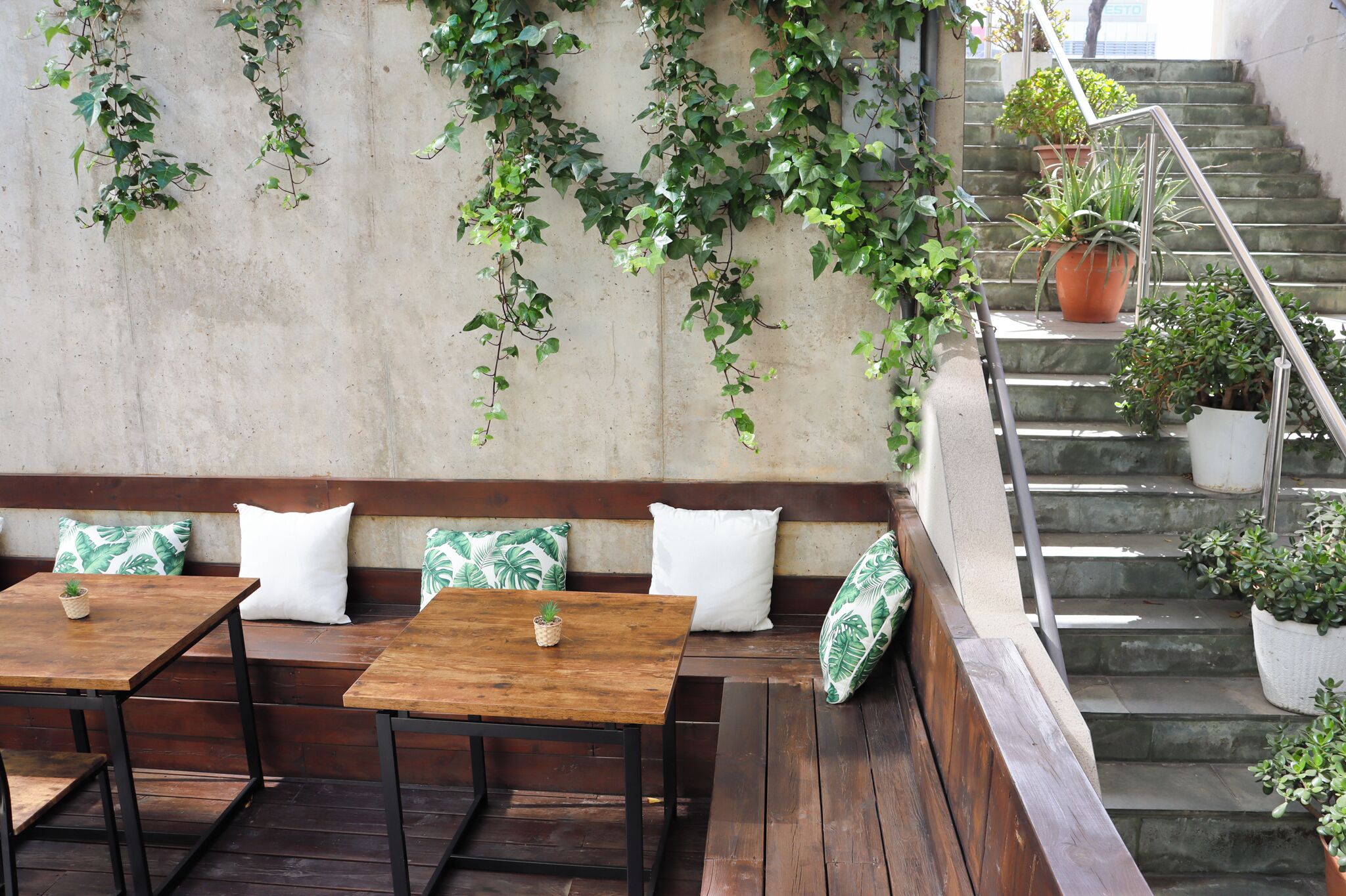Un área de estar al aire libre con bancos y mesas de madera, rodeada de plantas en macetas y una escalera.