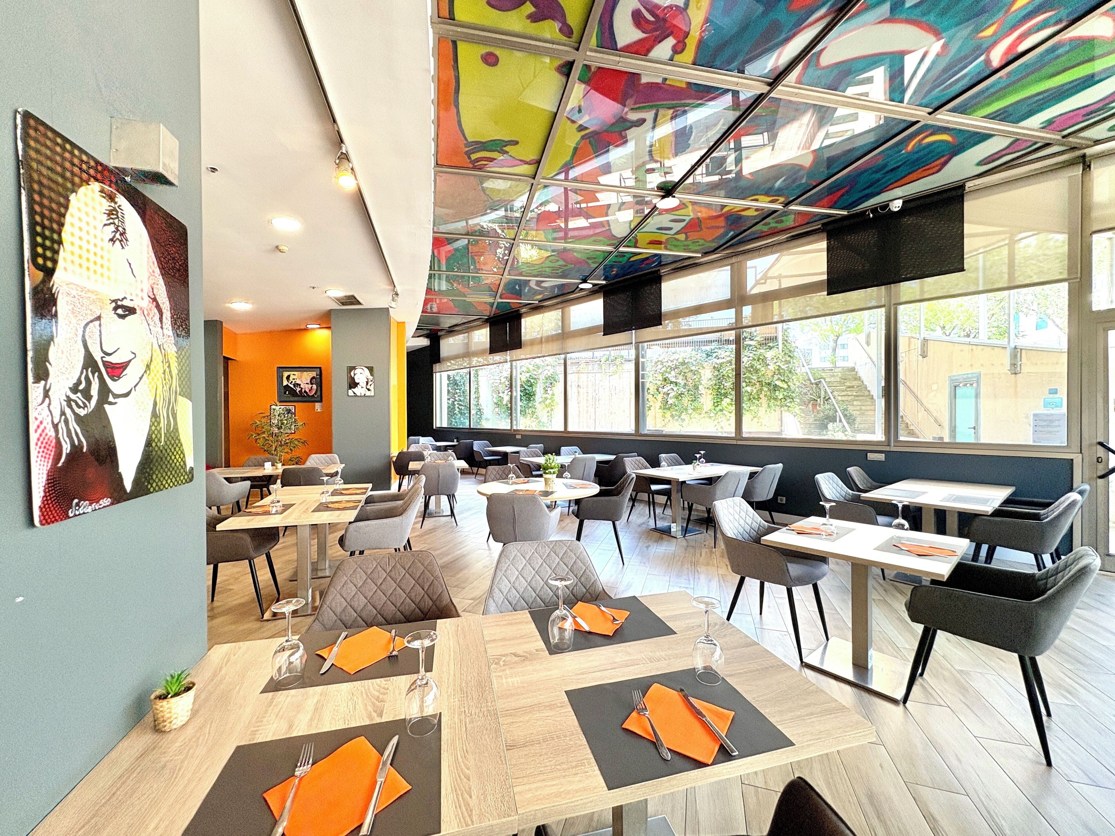 A modern restaurant with large windows, colorful ceiling art, and neatly arranged tables with orange placemats.
