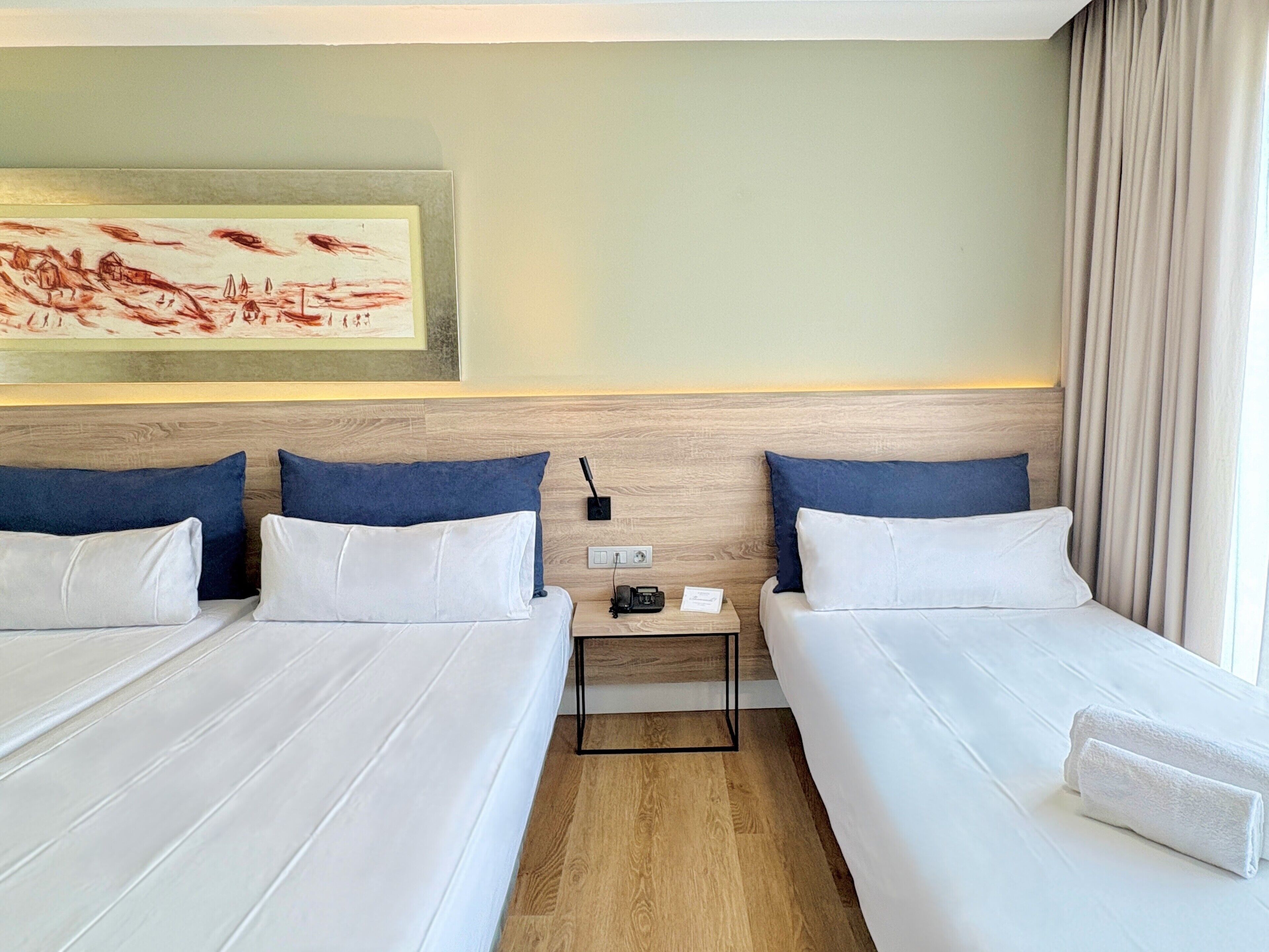 Una habitación de hotel con dos camas, una mesita de noche, un cuadro y una ventana con cortinas.