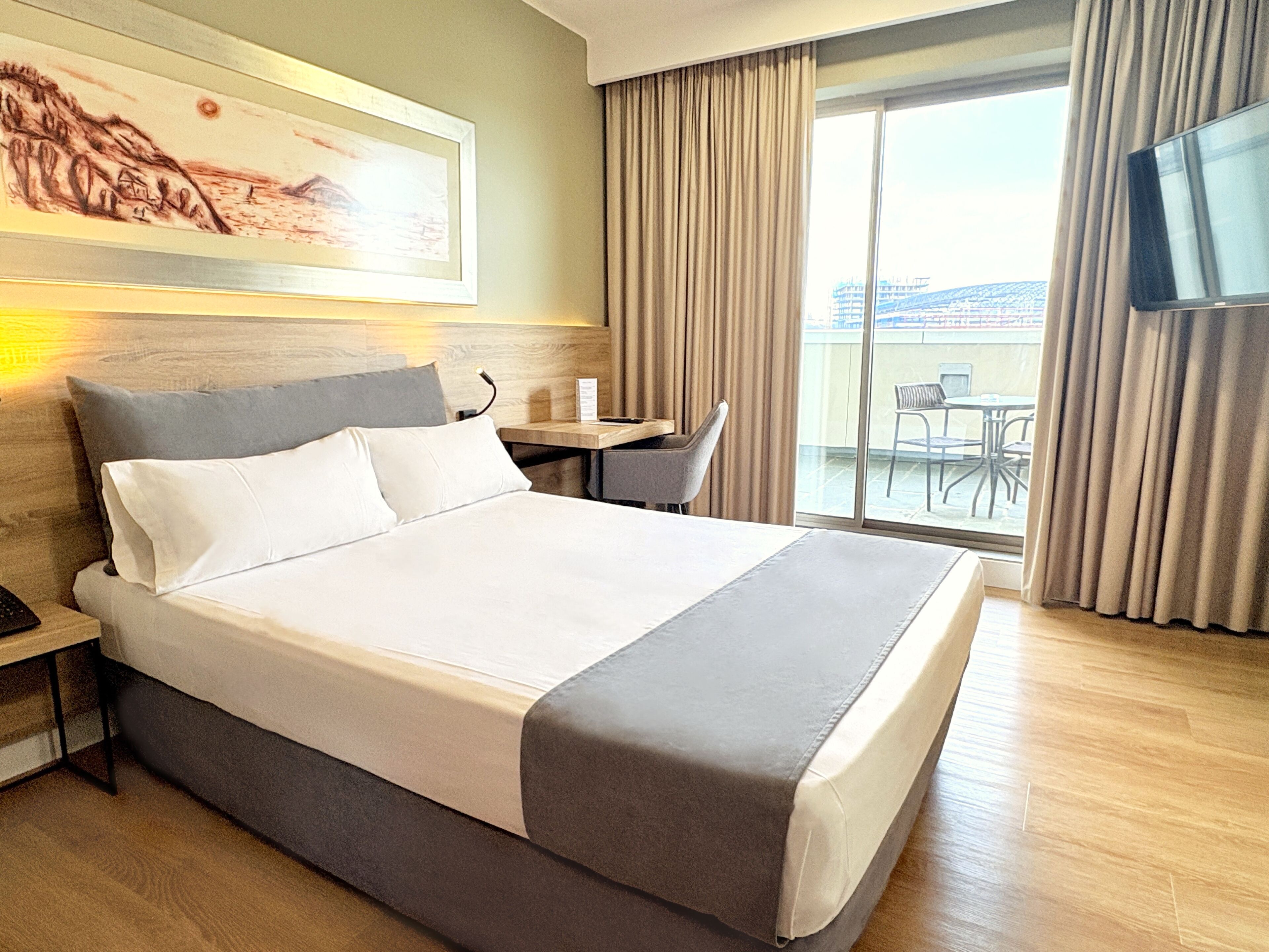 A hotel room with a large bed, a desk, a chair, a TV, and a balcony with a view.