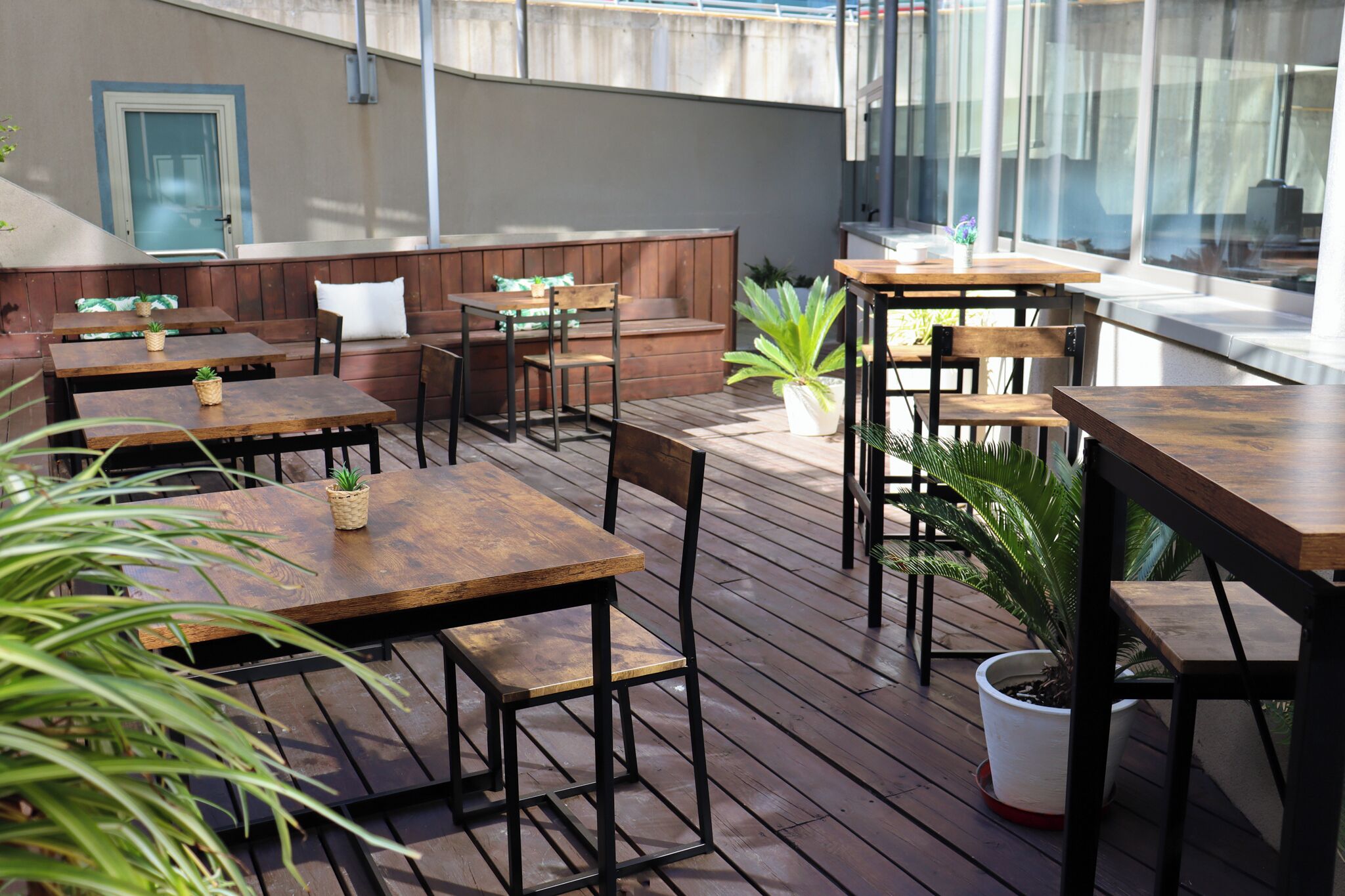 Une terrasse extérieure avec des tables et des chaises en bois, un coin salon avec des coussins et des plantes en pot.