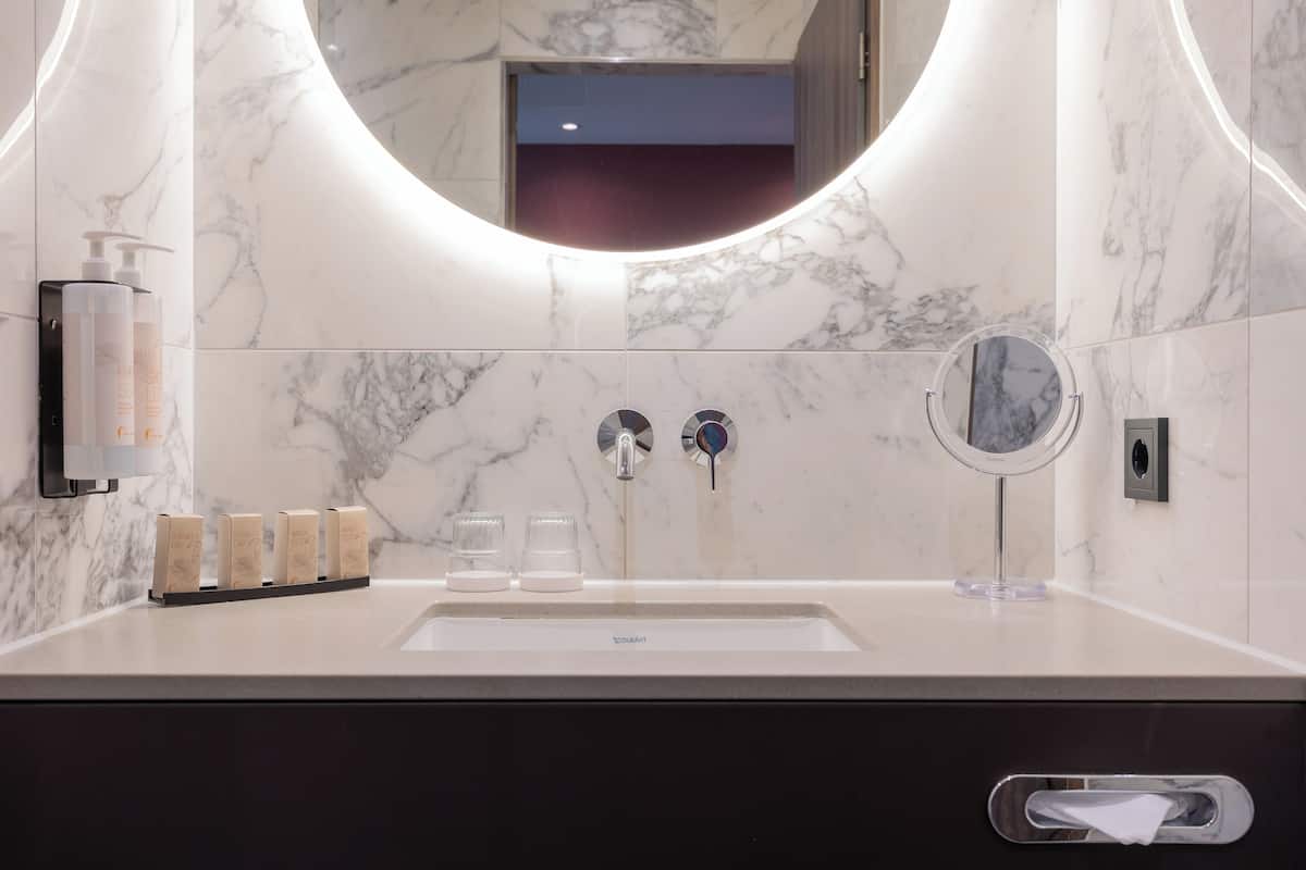 A modern bathroom with a marble sink, two faucets, a round mirror, and a soap dispenser.